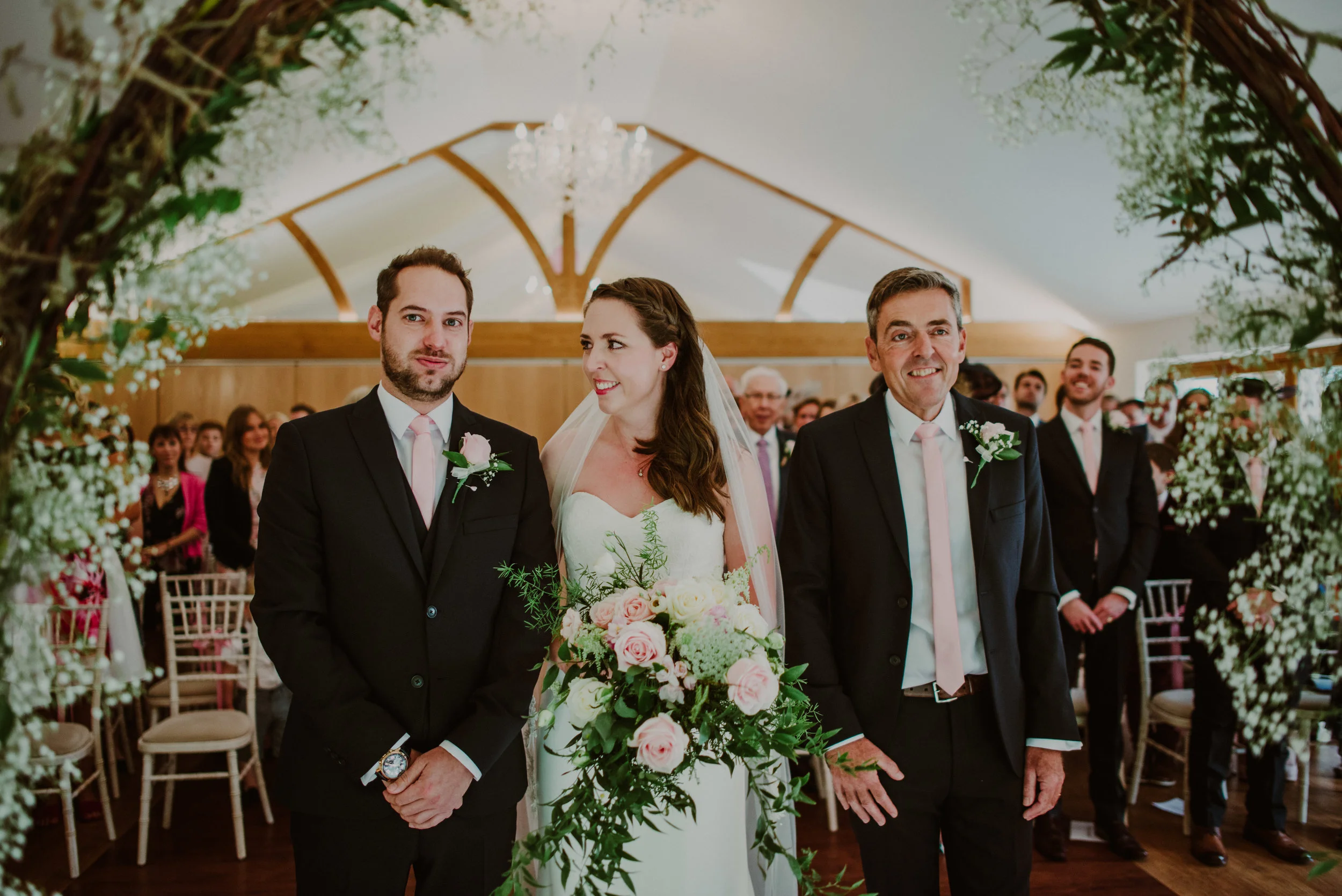 A bride standing between two men during a wedding ceremony, in a decorated indoor venue with guests watching in the background.