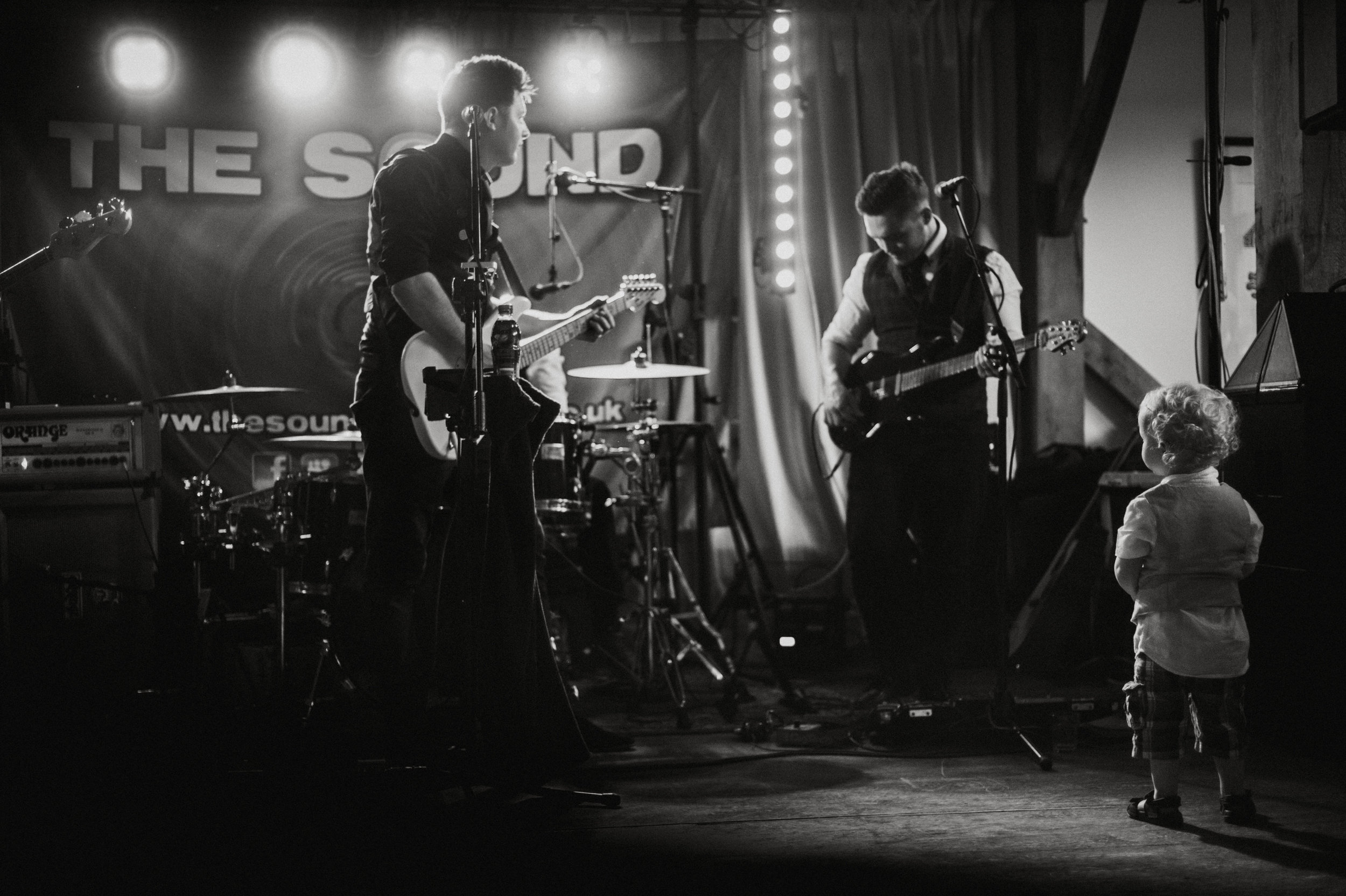 A black and white photo of a musical performance on stage. Two male musicians are playing guitars, with a drum set behind them. A young child stands in front of them, facing the stage, watching the performers. The background shows a banner reading 'T