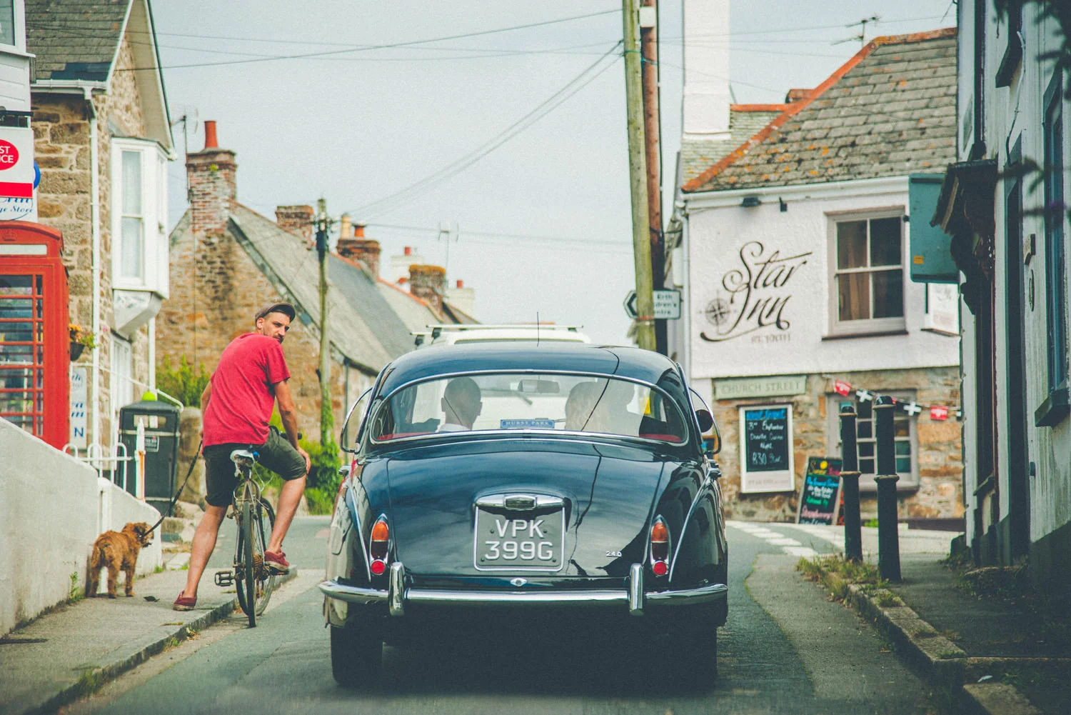 A man in a red shirt and black shorts sitting on a bicycle with a small brown dog on a leash walking beside him on a street lined with stone buildings. A vintage black car with the license plate 'VPK 399G' is parked in front of him, and there are sig