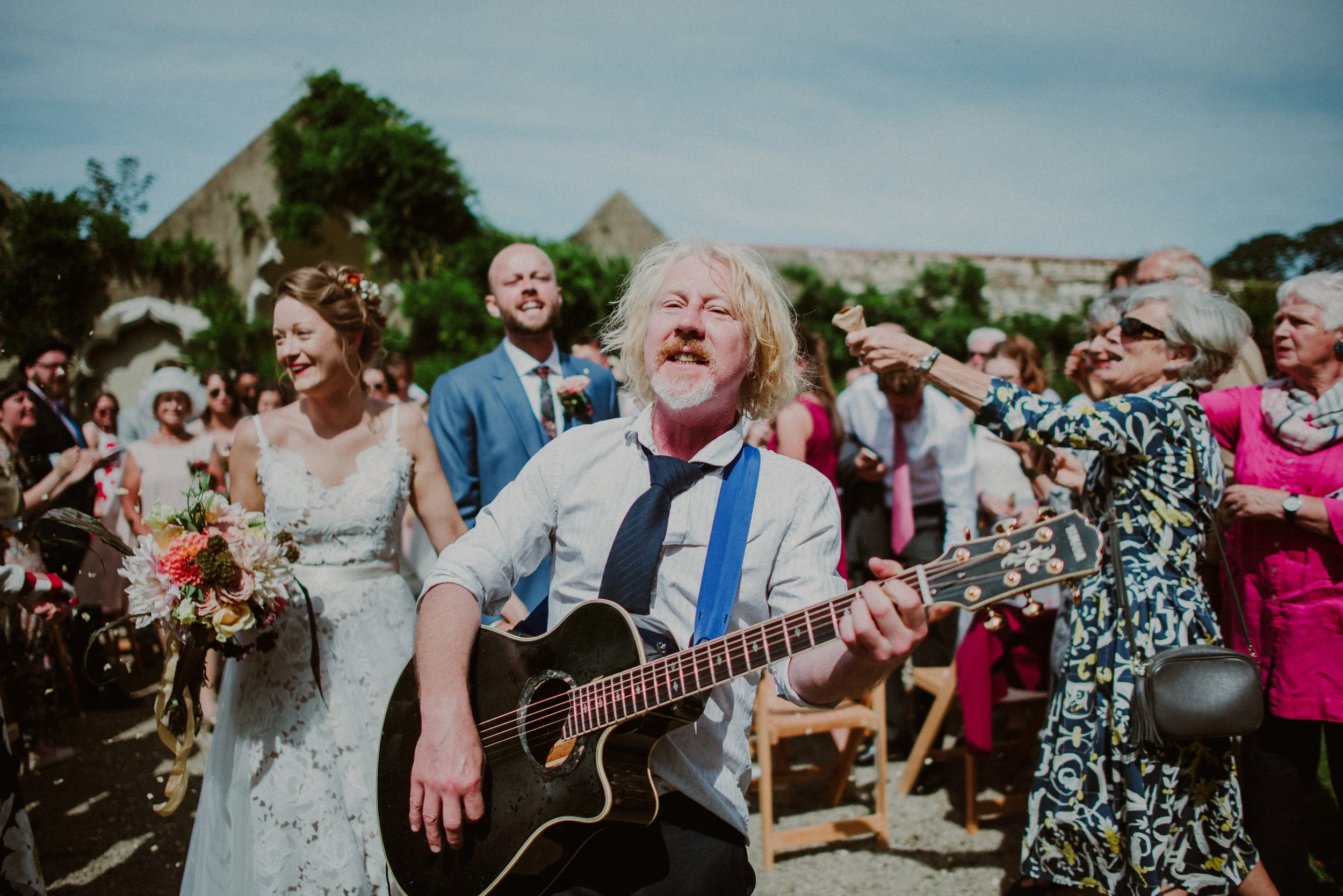 Man with long blond hair playing guitar at outdoor wedding, surrounded by smiling guests, including bride in white dress holding bouquet and groom in blue suit, in sunny setting with greenery and stone buildings in background.