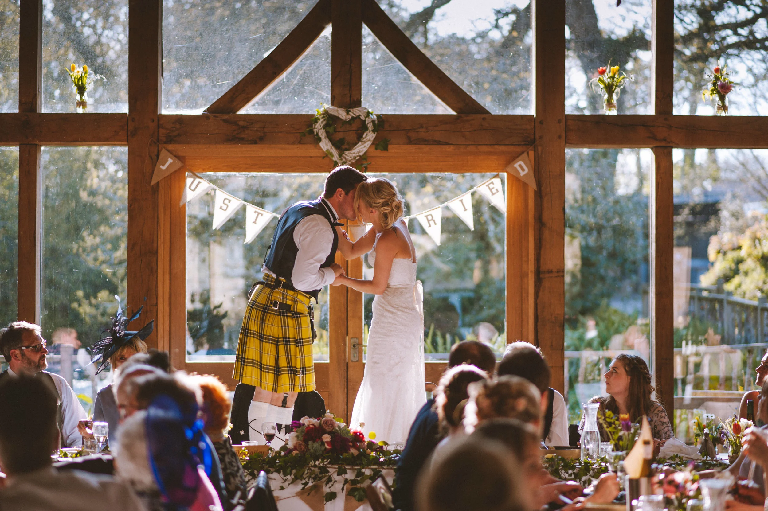 A bride and groom are dancing on their wedding reception table, touching foreheads. The groom is wearing a traditional Scottish kilt, and the bride is in a white wedding dress. Guests are seated around the table, watching them, with a star-shaped ban