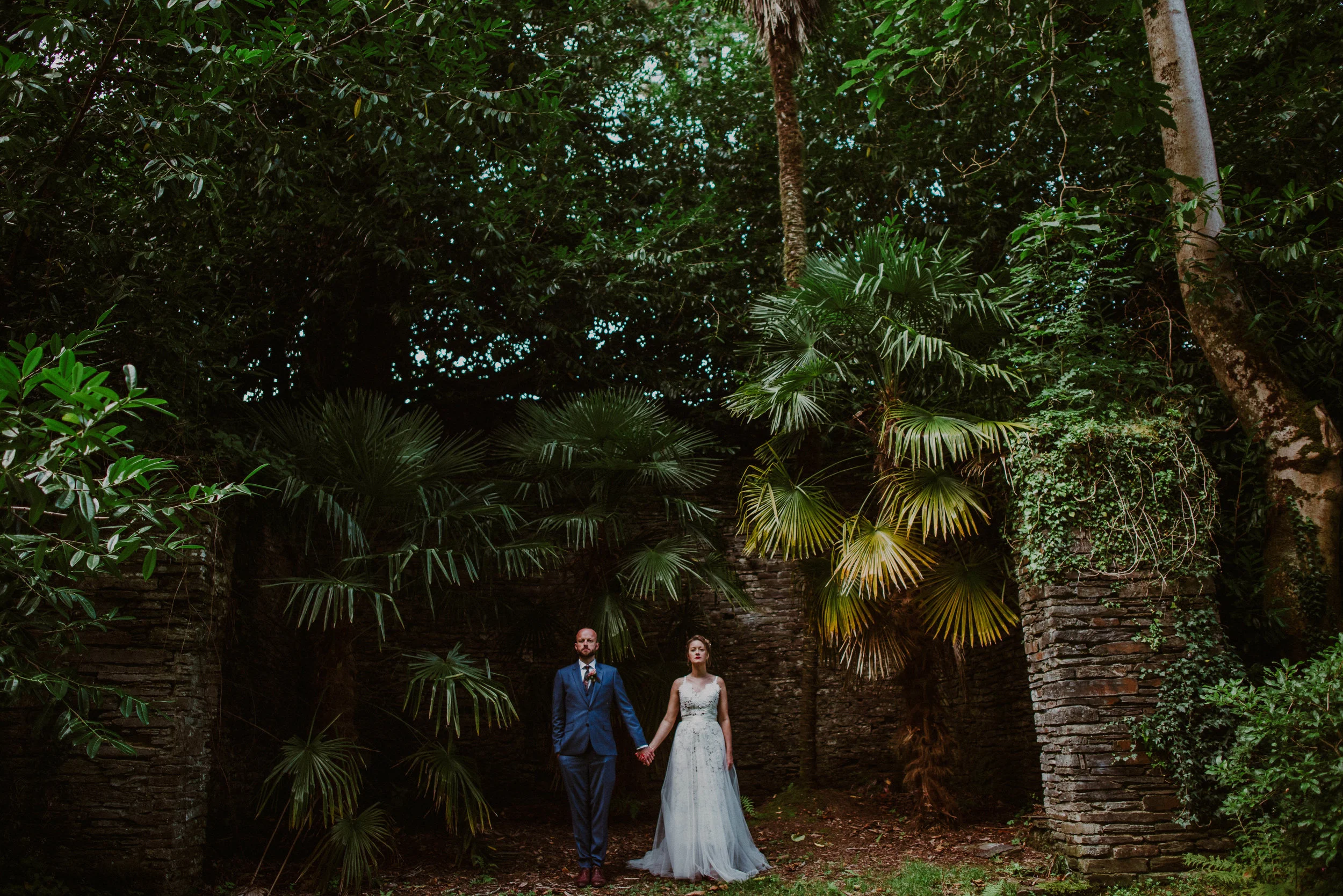 A bride and groom holding hands stand in a lush, green outdoor setting with dense foliage and stone pillars.