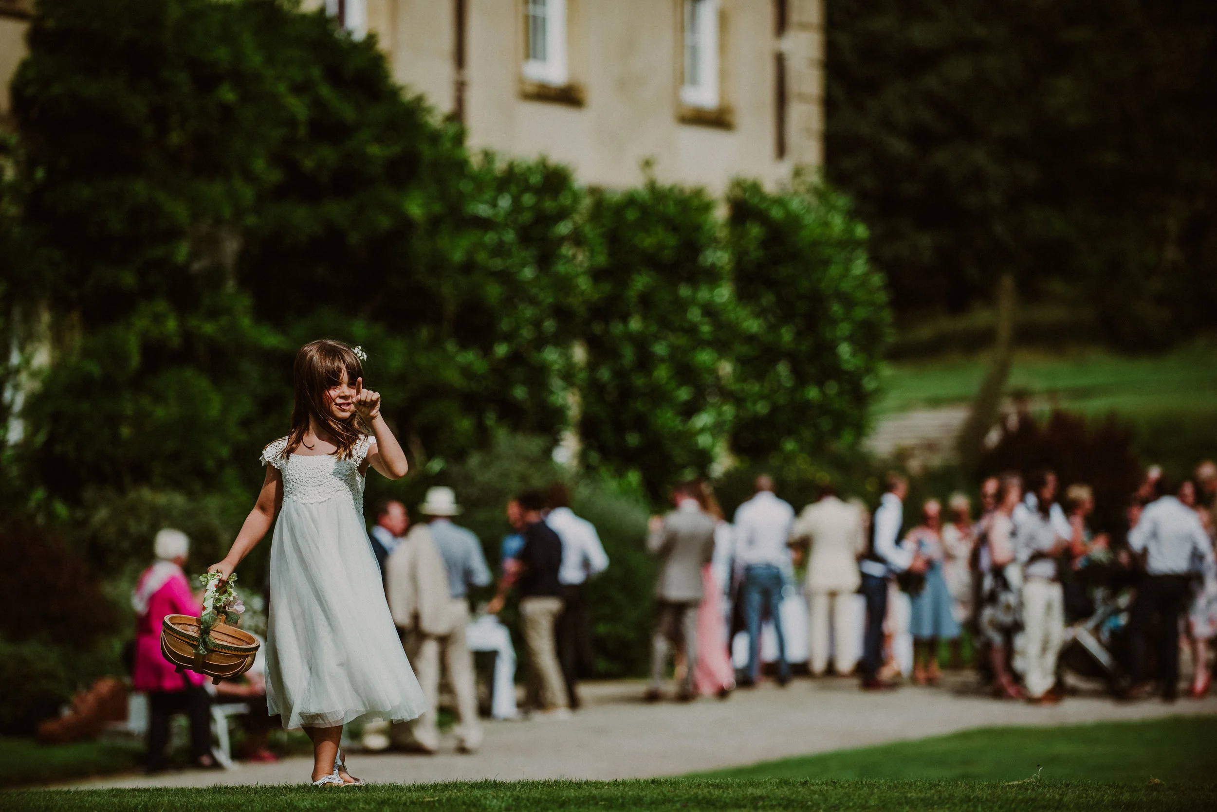 A young girl in a white dress standing on a lawn, with a crowd of people in formal attire gathered in the background, outdoors during daytime.