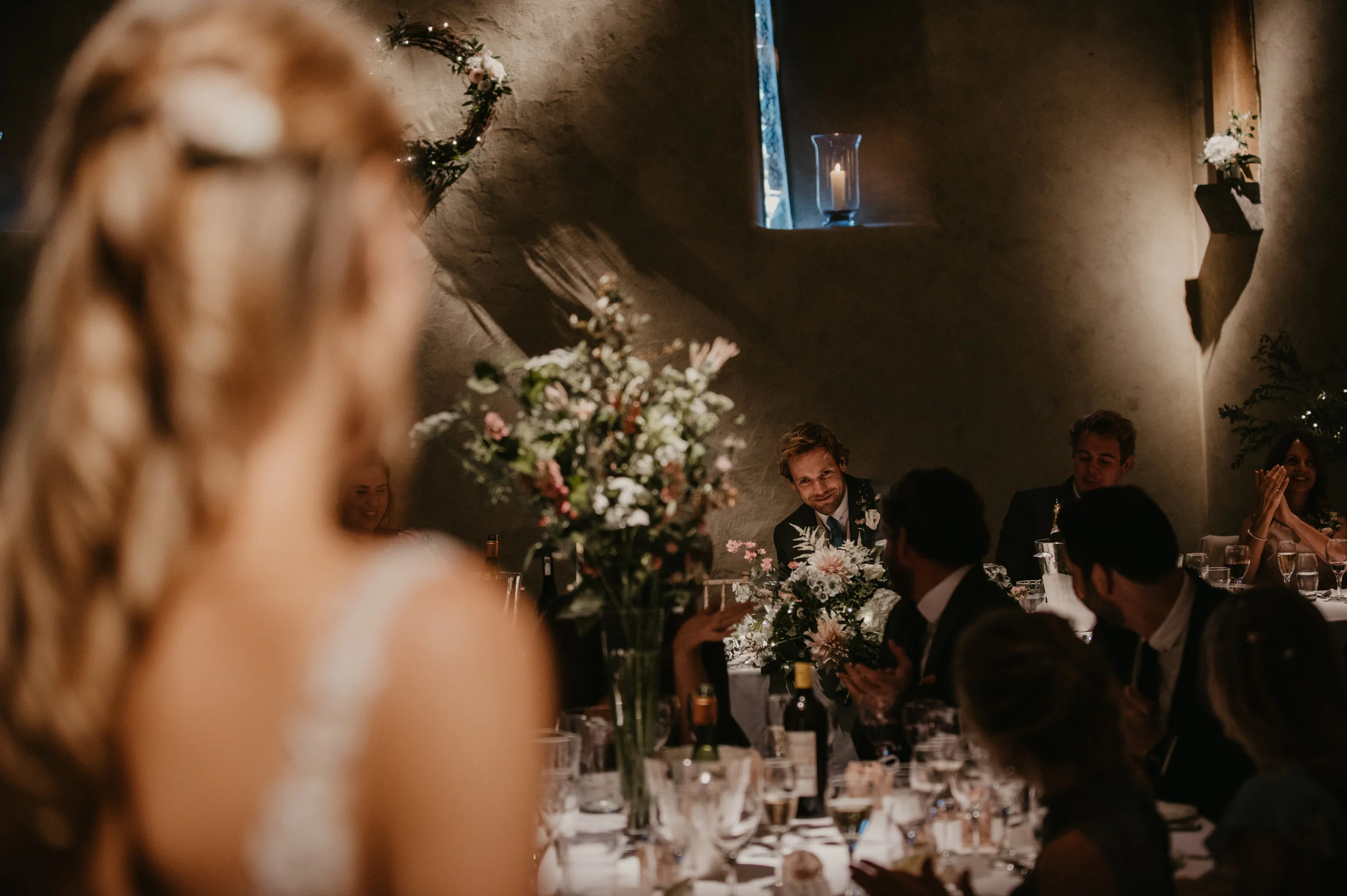 People at a wedding reception, with a woman in a wedding dress in the foreground and guests, including a man in a tuxedo, sitting around a decorated table with flowers and drinks. The room has a small window with a lit candle on the sill.