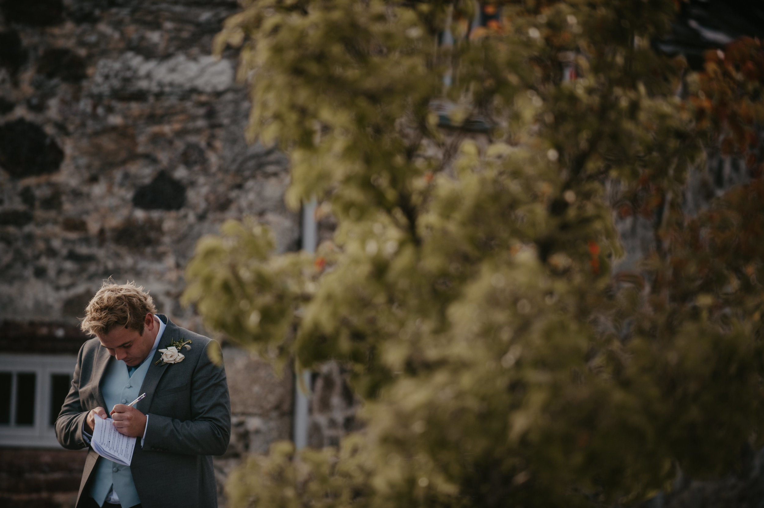 A man in a gray suit and a white tie with a white flower boutonniere, standing outdoors, writing on a small notepad or piece of paper, with a stone building and green trees in the background.