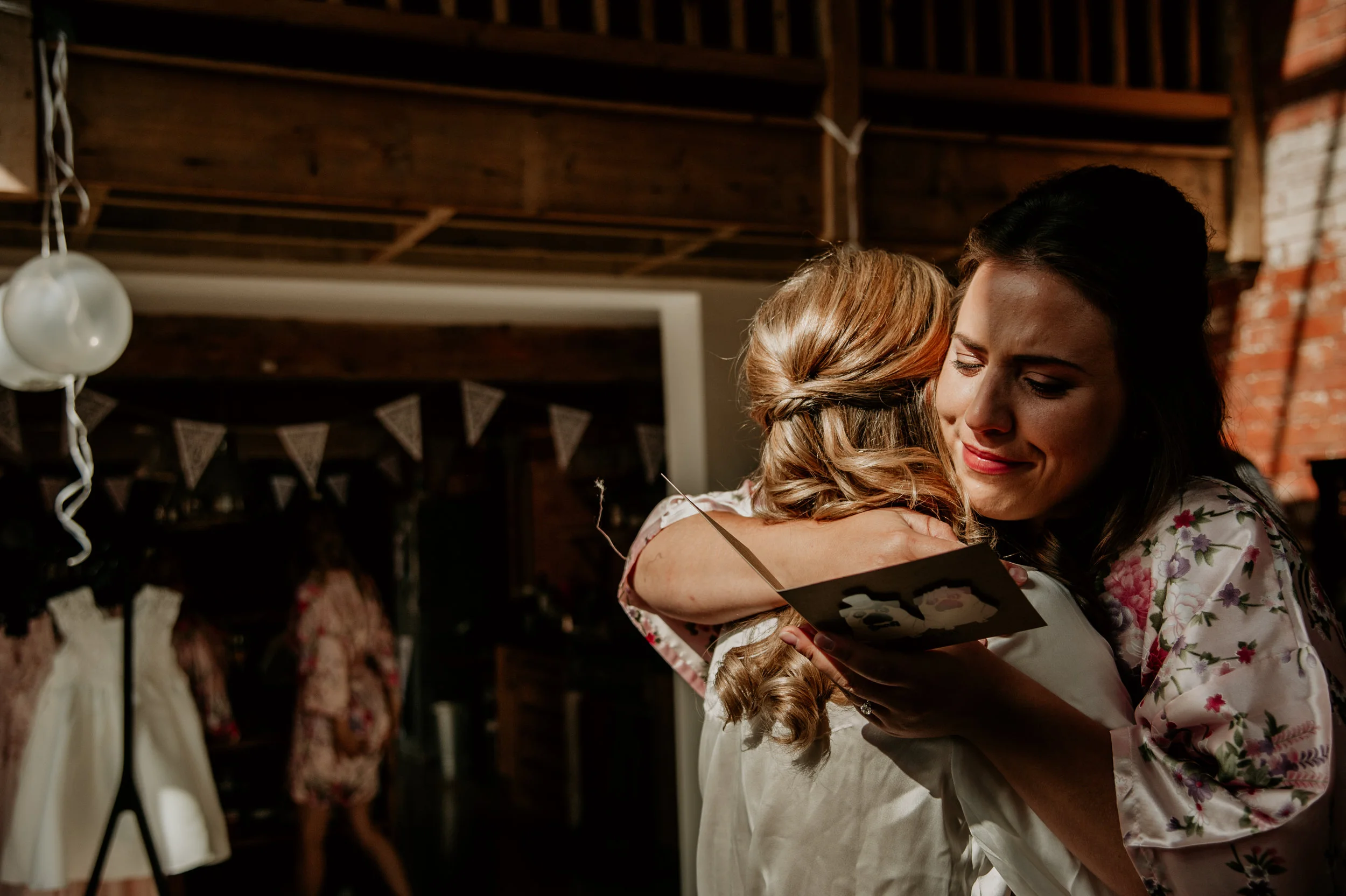 Two women hugging, one holding a greeting card, in a rustic indoor setting with wedding dresses and decorations in the background.
