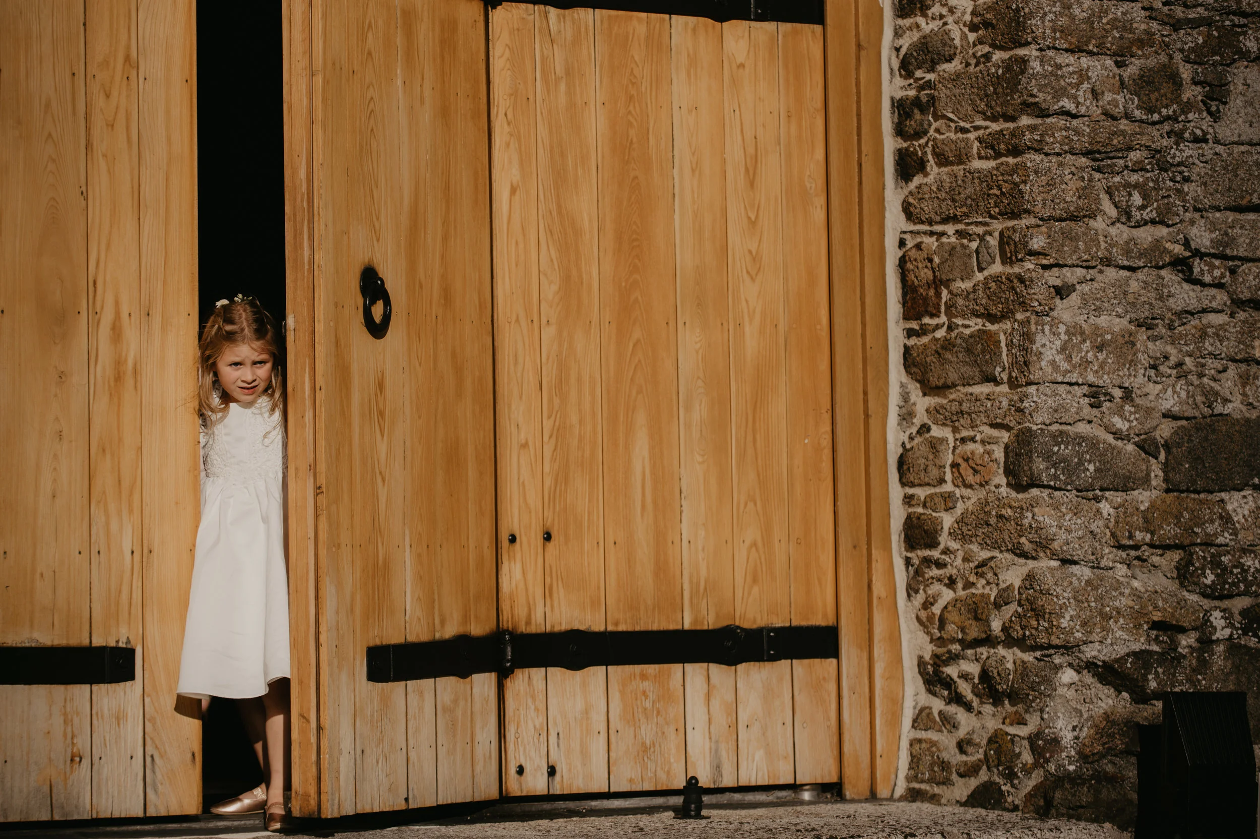 A young girl in a white dress and brown shoes looks out from behind a large wooden door, with a stone wall on the right side of the door.