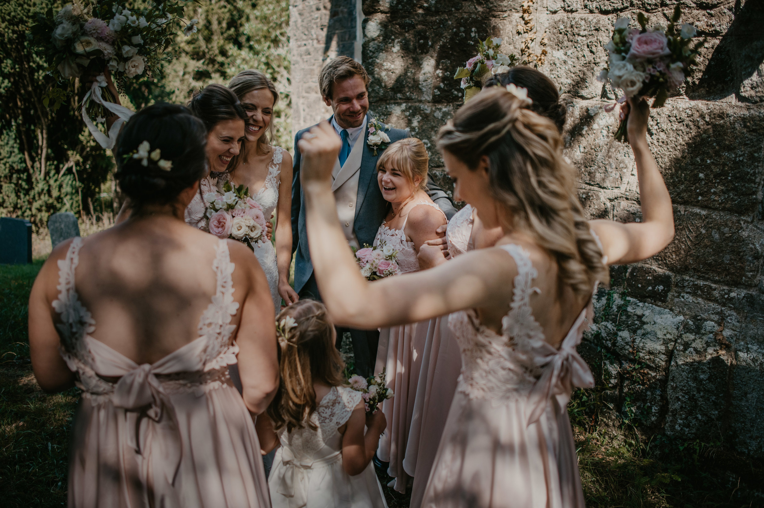 Group of people, including bridesmaids and a bride, celebrating outdoors at a wedding, smiling and raising their arms in excitement.