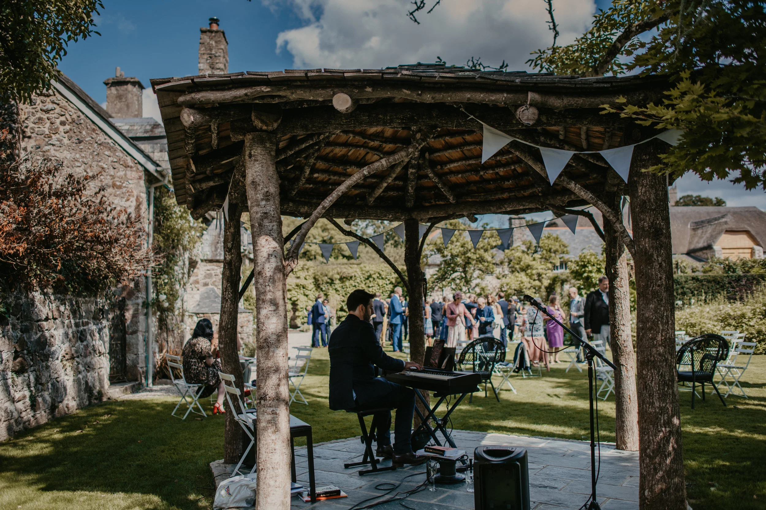 A musician playing a keyboard under a rustic wooden gazebo at an outdoor garden party with guests mingling in the background.