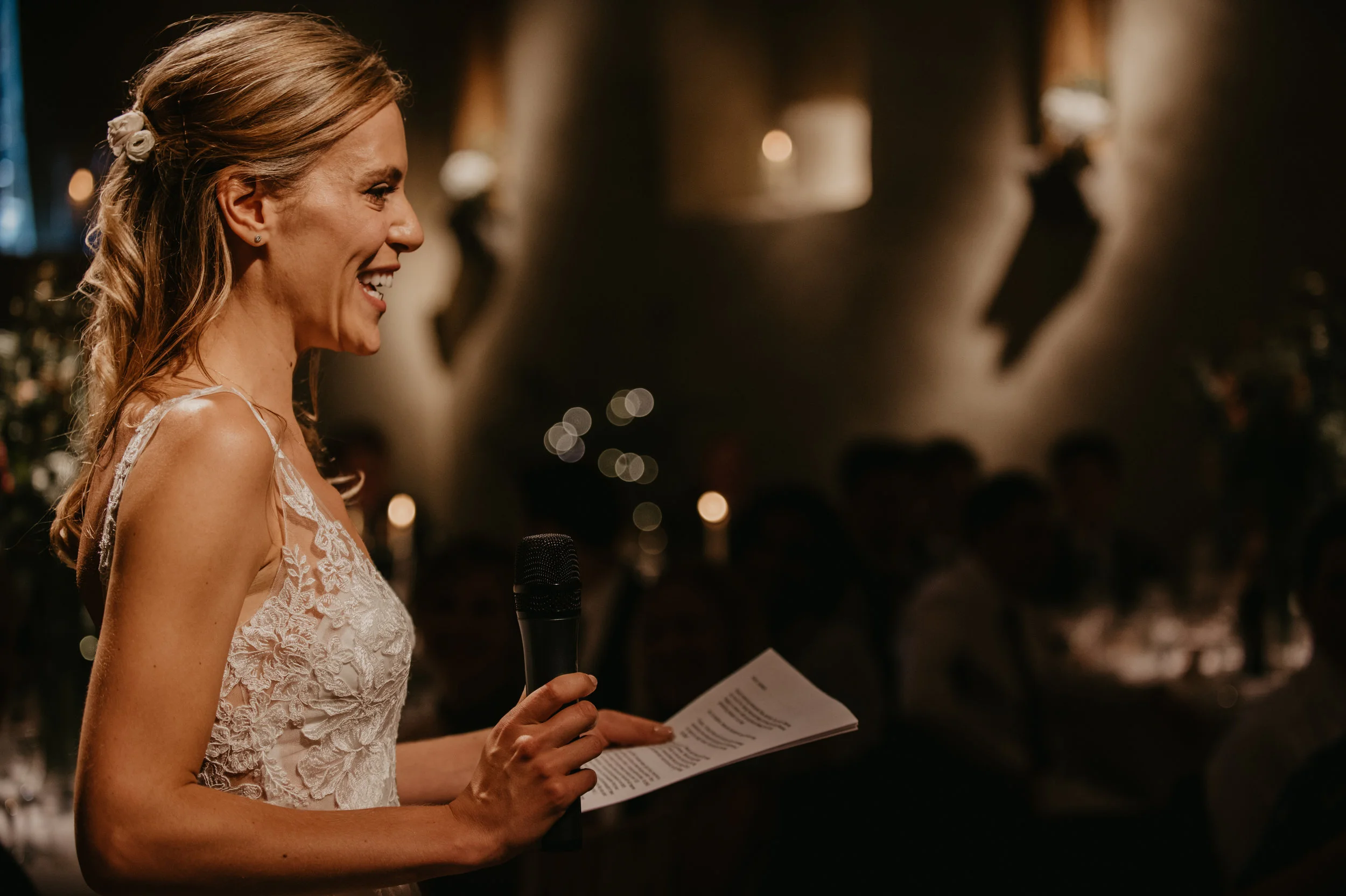 A woman in a lace dress is giving a speech at a wedding reception, holding a microphone and a paper, smiling and facing the audience in a dimly lit room.