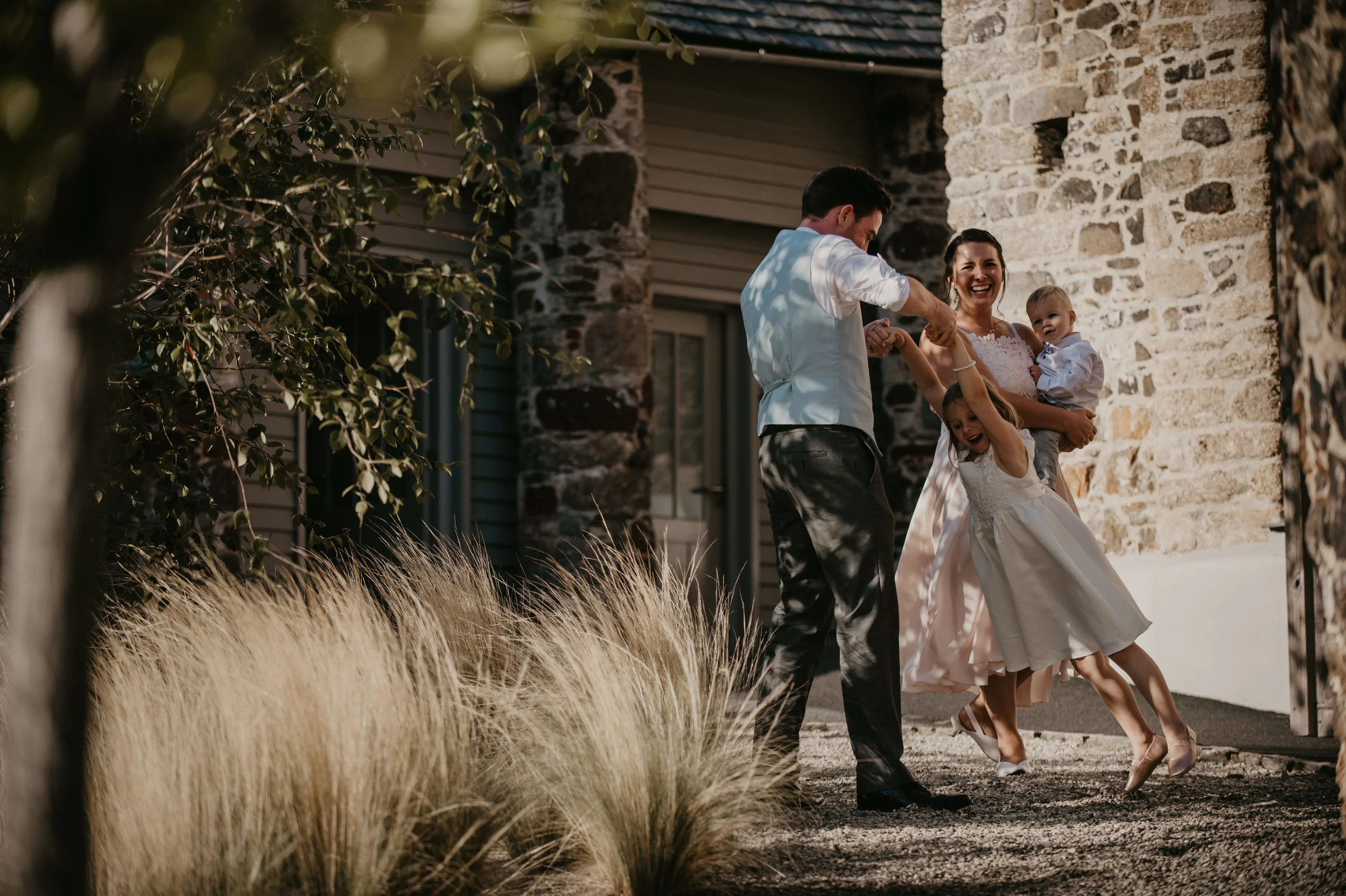 A family dancing outside near a stone wall, with two children, on a sunny day.