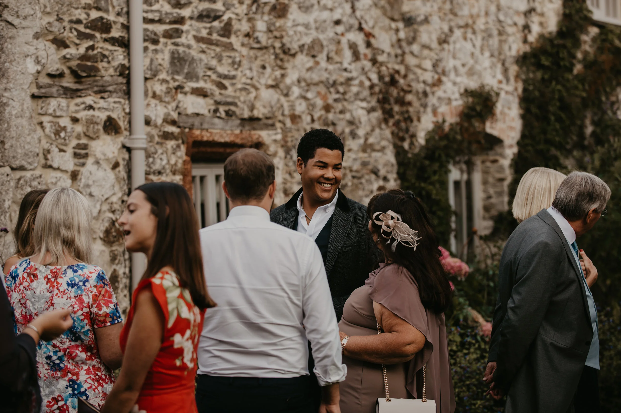 Group of people socializing outdoors in front of a stone wall, including a smiling man at the center wearing a dark blazer, surrounded by women and men dressed in semi-formal attire.