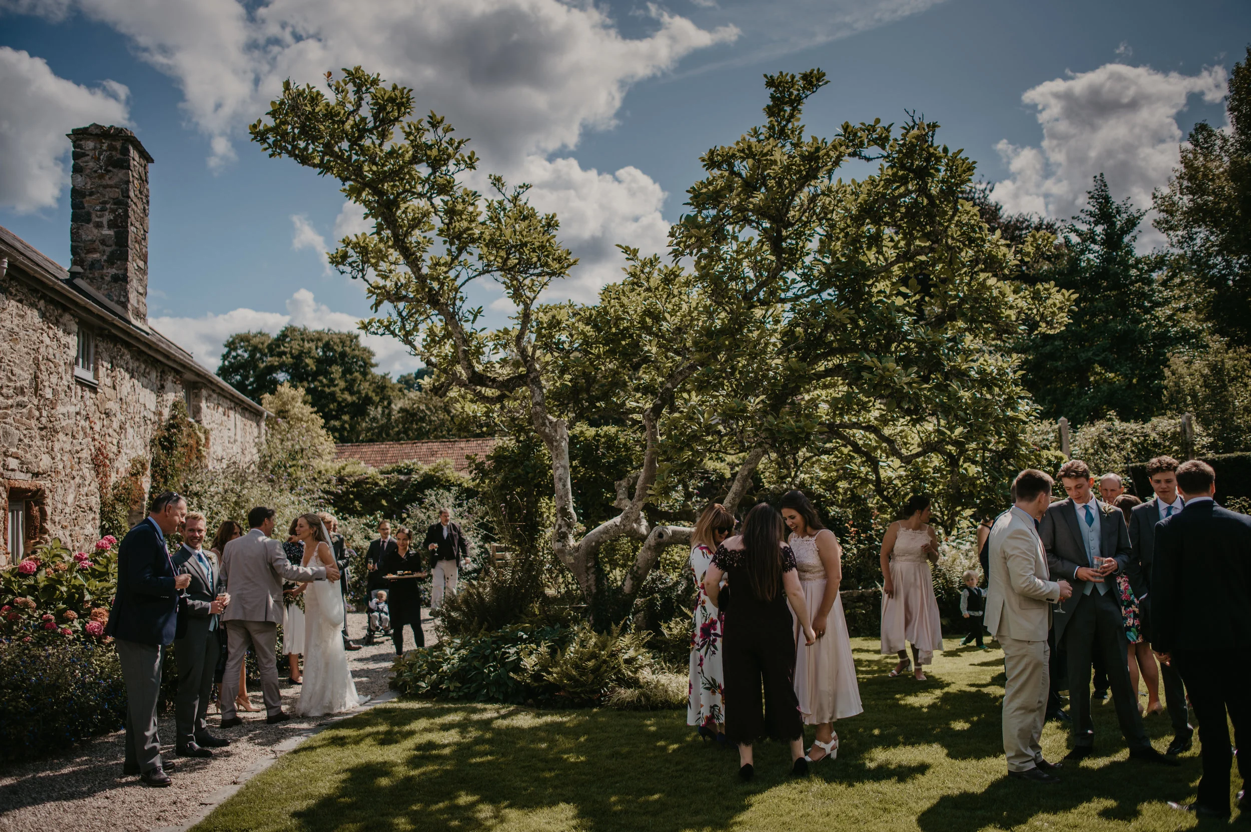 People gathering in a garden on a sunny day, dressed in semi-formal attire, with a stone building, trees, and shrubs in the background.