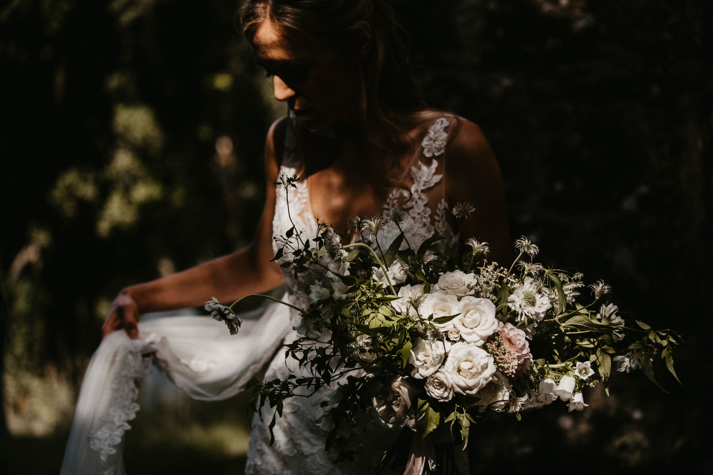 A woman holding a large bouquet of white flowers, wearing a white dress with lace details, outdoors in a shaded area with sunlight filtering through trees.