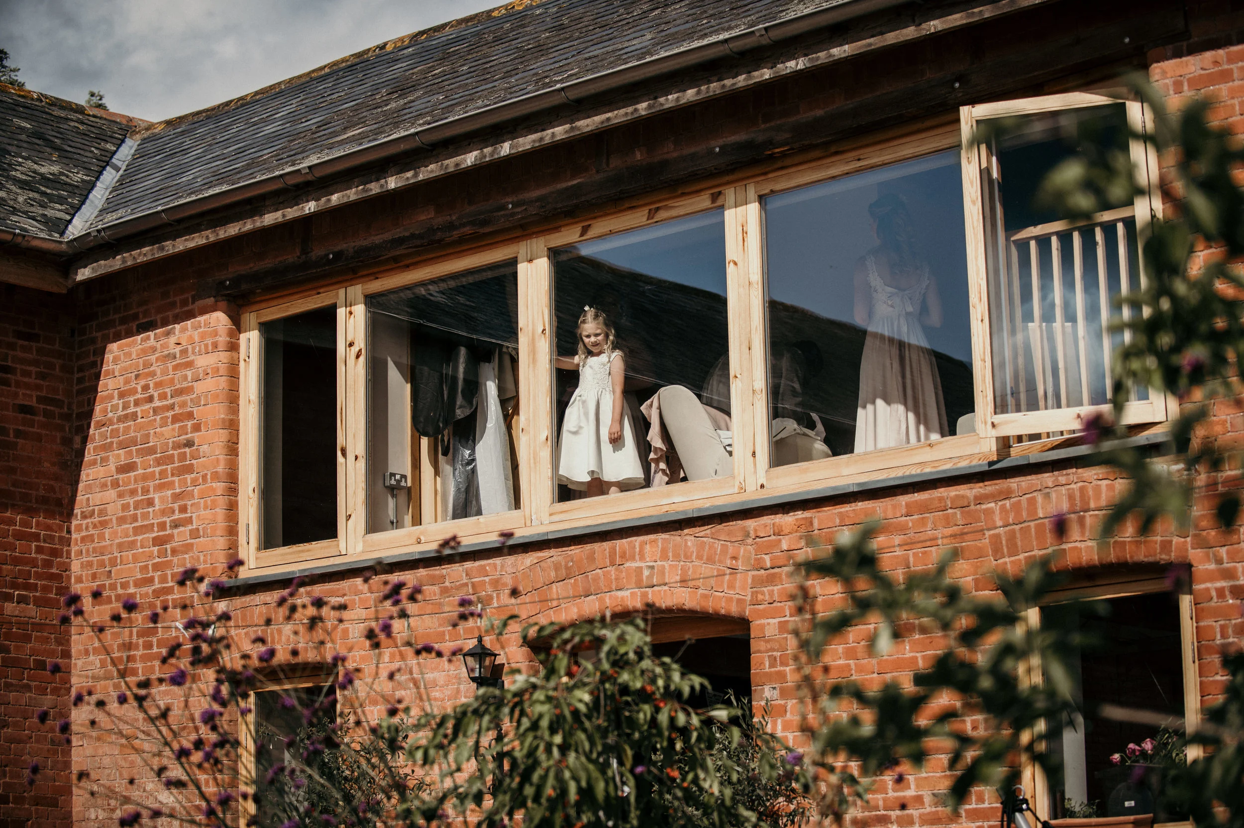 Girl in a white dress standing inside an unfinished brick house, looking out a large window, with clothes hanging inside