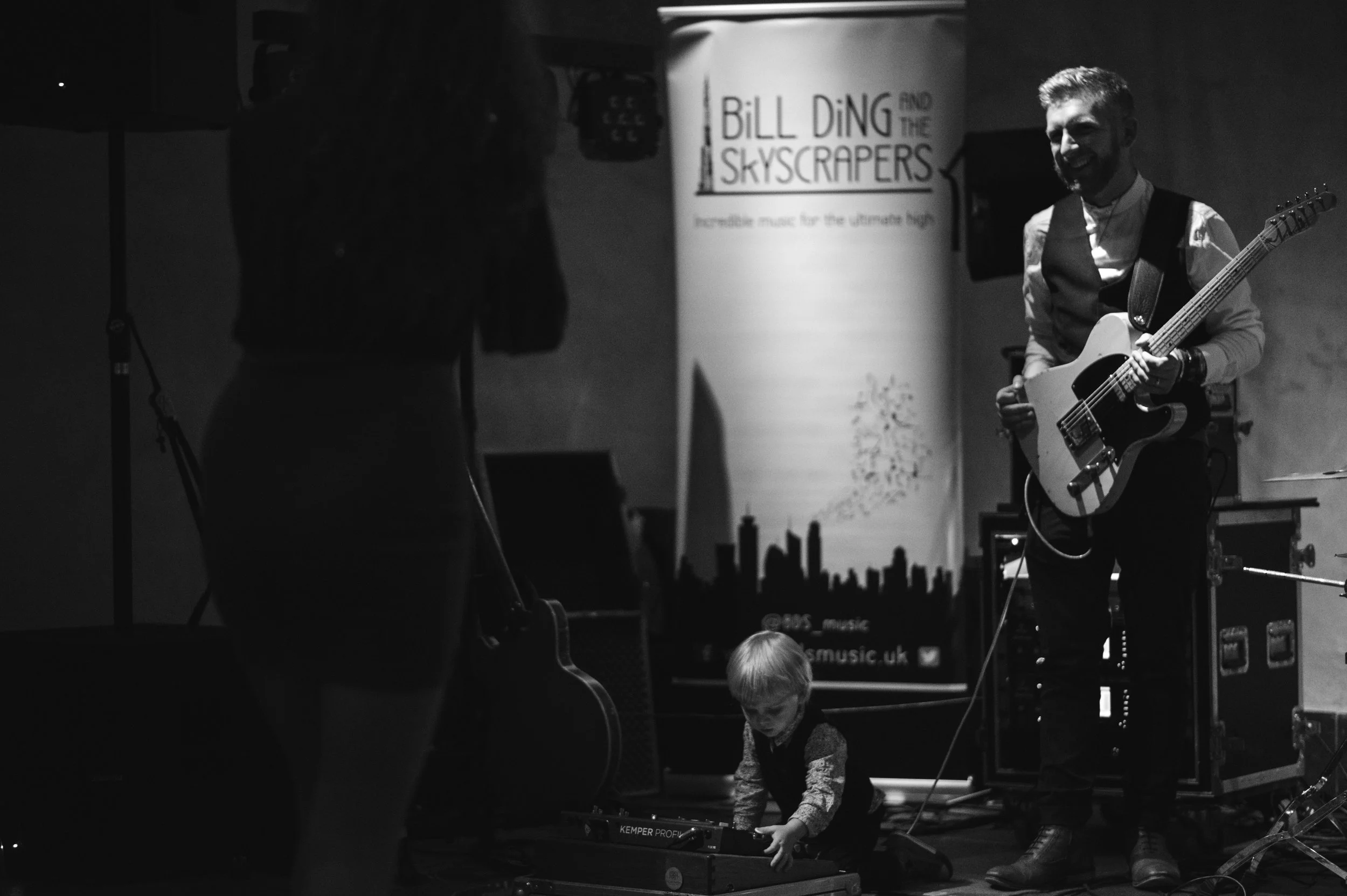 A black and white photo of a young child playing a keyboard on stage with musicians. An adult man with a beard, dressed in a vest and shirt, plays an electric guitar and smiles. In the background, a banner reads 'Bill Ding and the Skyscrapers.'