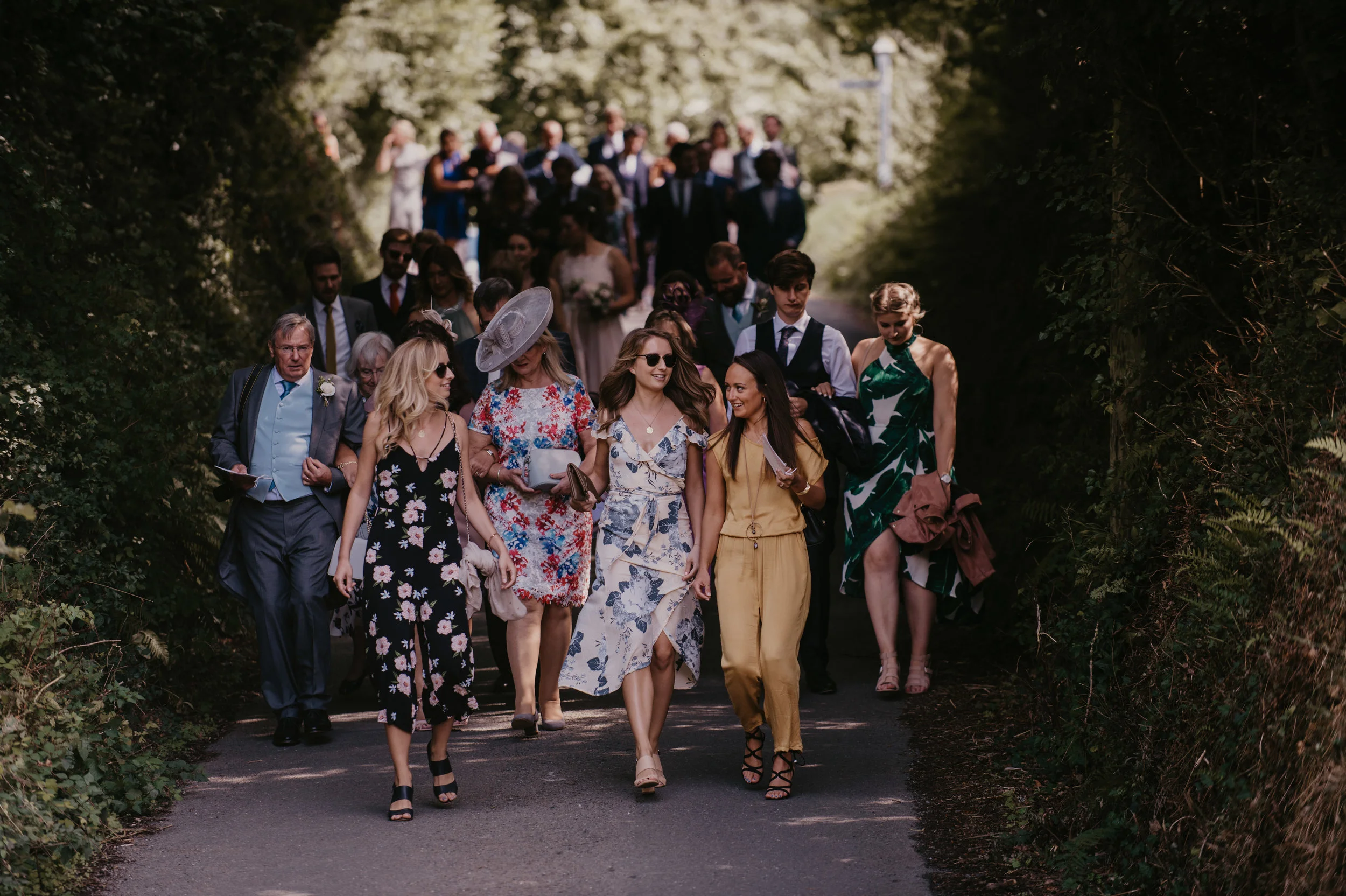 People attending a wedding, walking down an outdoor path surrounded by greenery, with some dressed in formal and semi-formal attire.