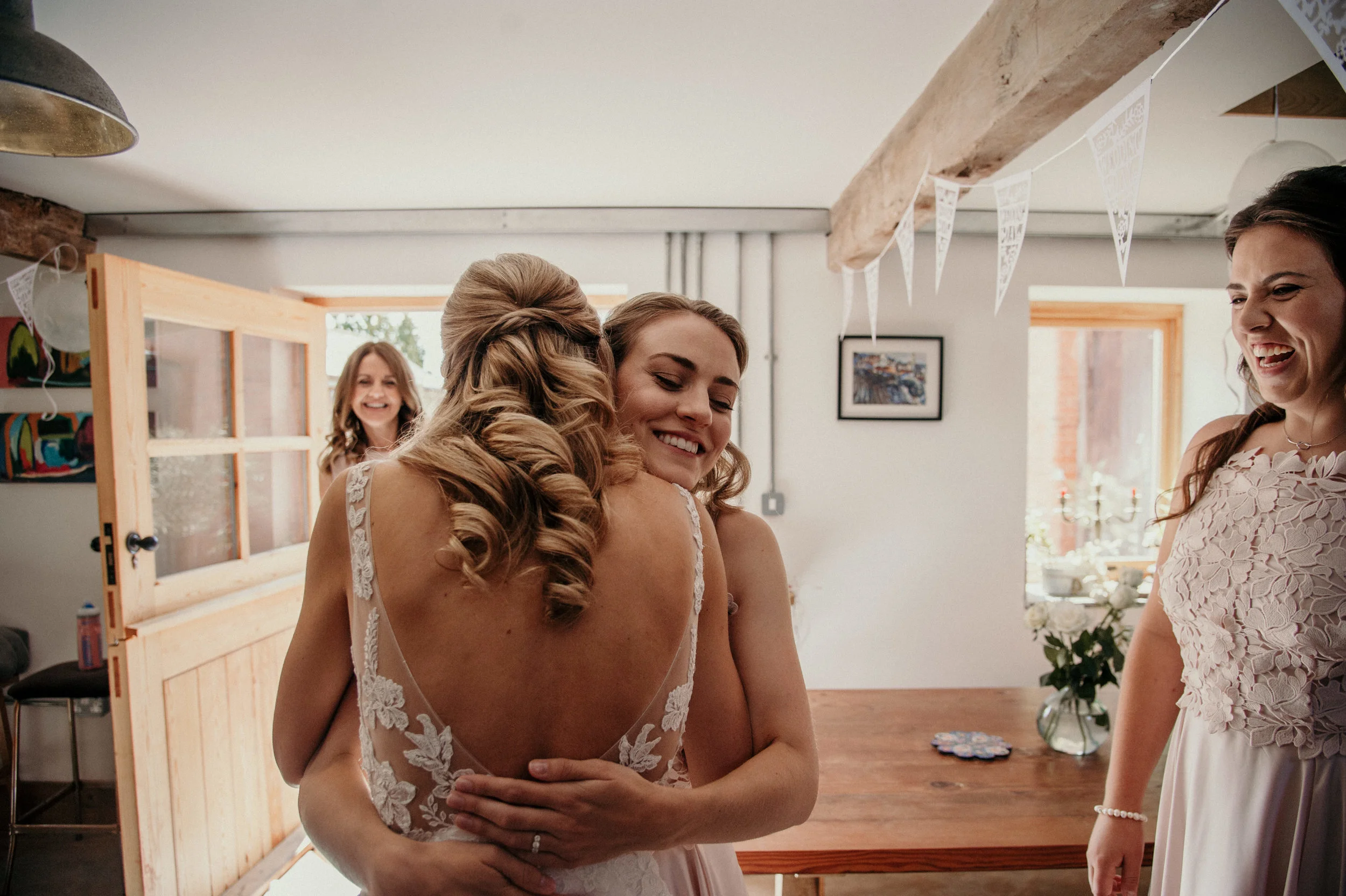 Two women embracing, one in a wedding dress, with two other women smiling nearby in a room decorated for a celebration.