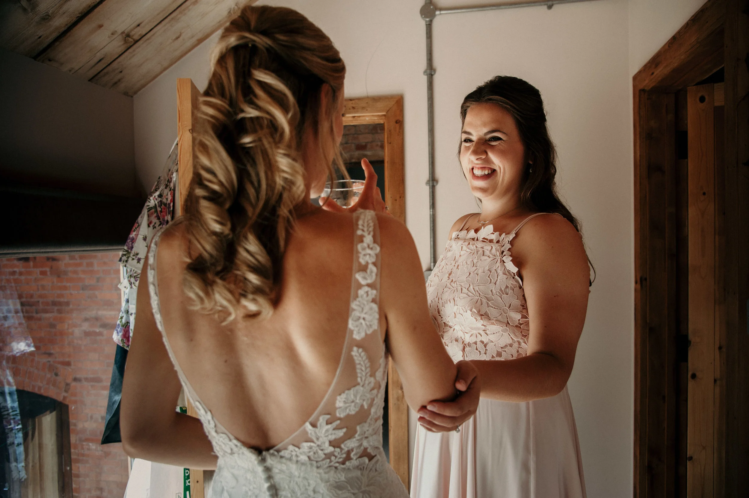 Two women in a warmly lit room, smiling and holding hands. One woman is wearing a wedding dress with lace details and open back, while the other is dressed in a light pink dress with floral lace top. They appear happy and excited, possibly during a w