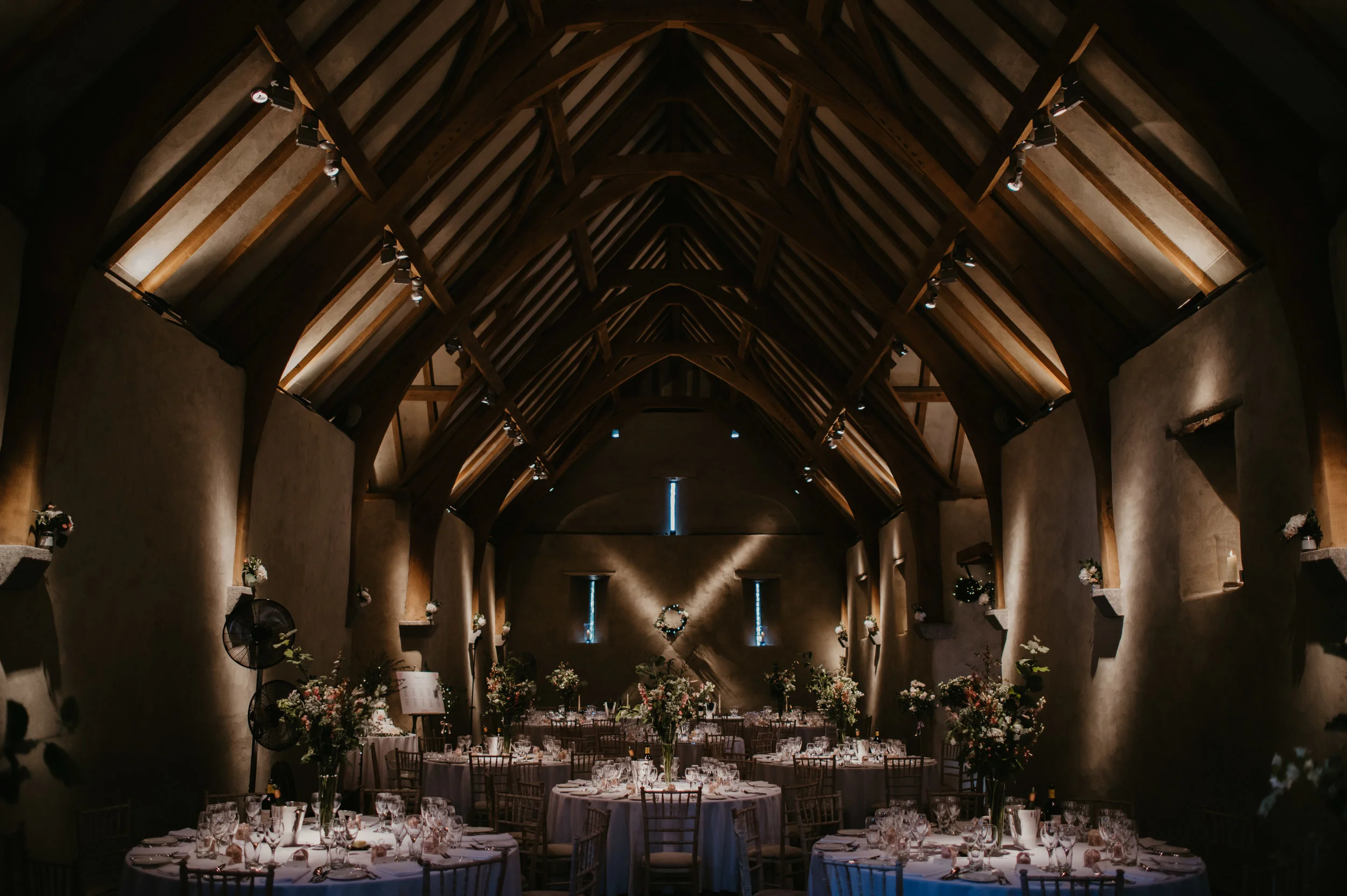 A decorated wedding or event reception inside a rustic barn with wooden beams, round tables with white tablecloths, floral centerpieces, and chairs arranged around the tables.