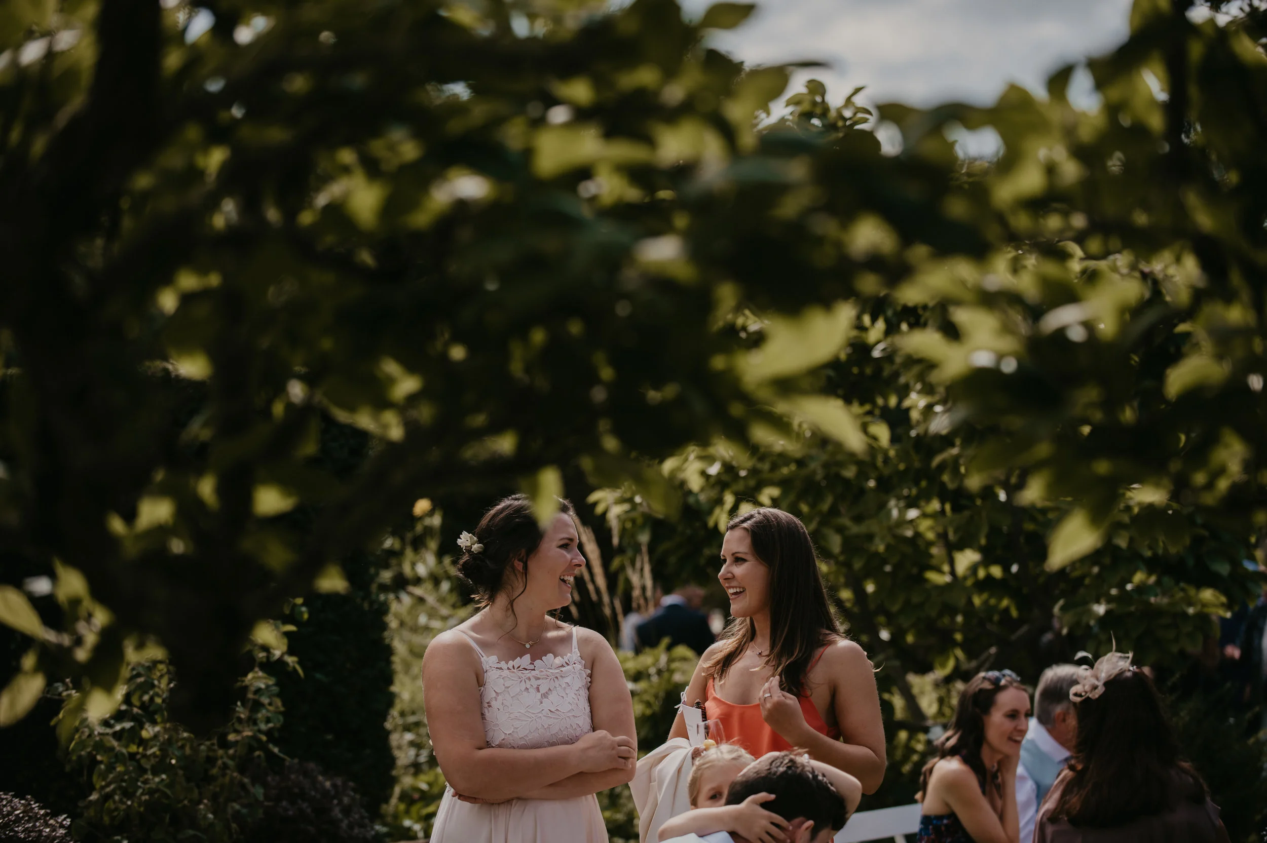 Two women are talking and smiling at a social event outdoors, surrounded by other people. They are under leafy green trees.