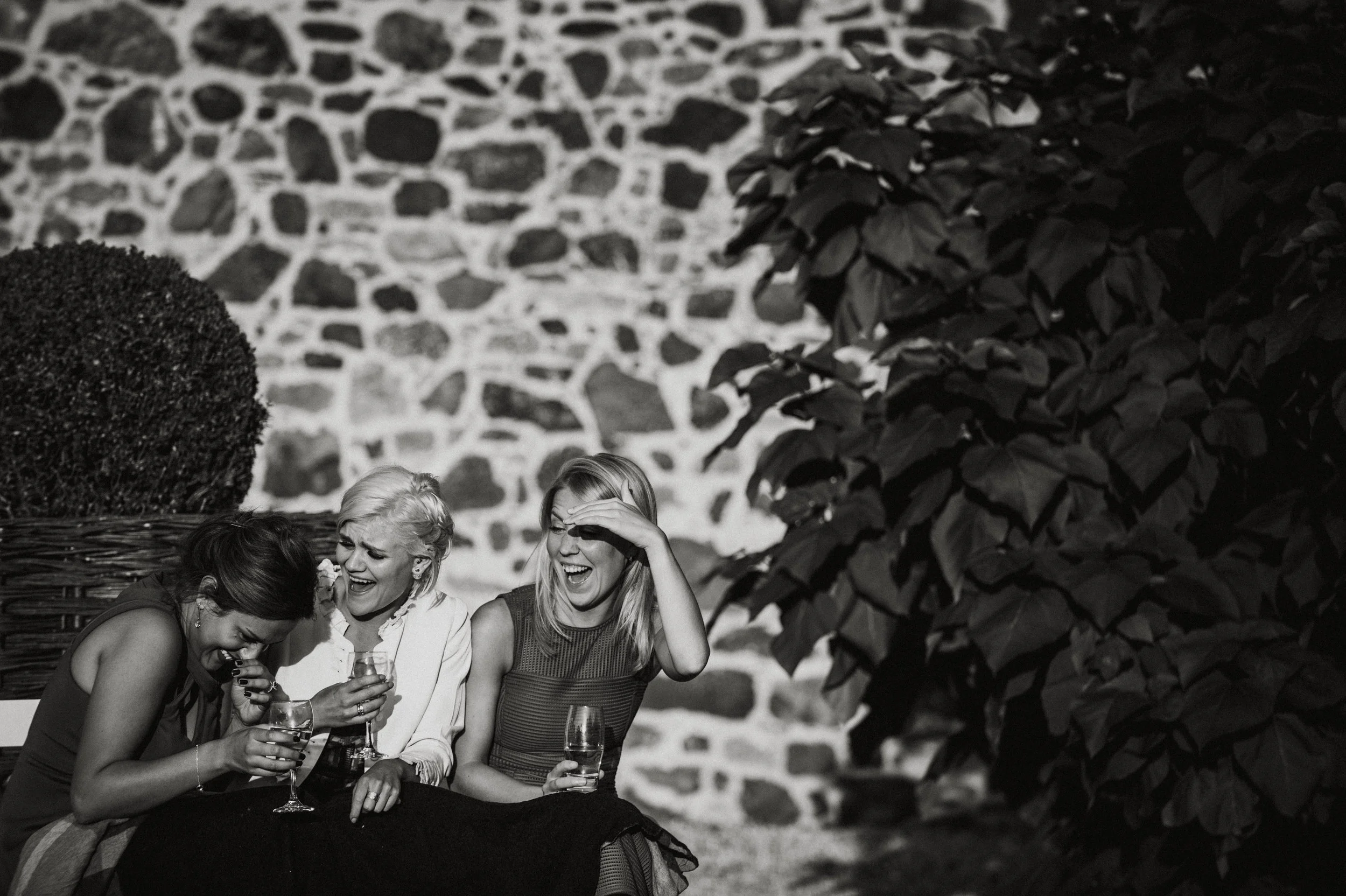 Three women sitting outdoors against a stone wall and large bush, laughing and holding drinks.