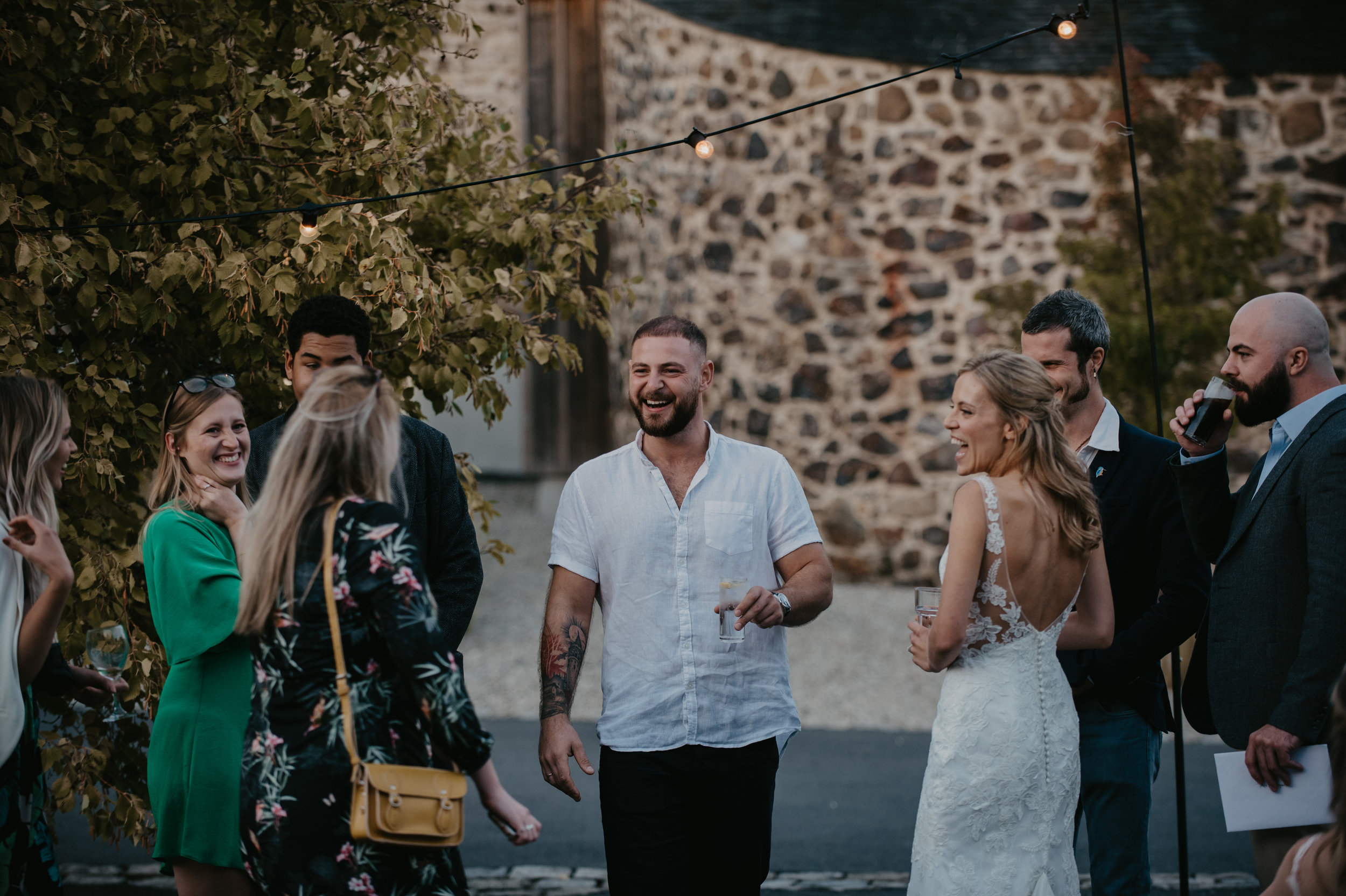 Group of people at an outdoor wedding reception, smiling and chatting, with string lights above and a stone wall in the background.