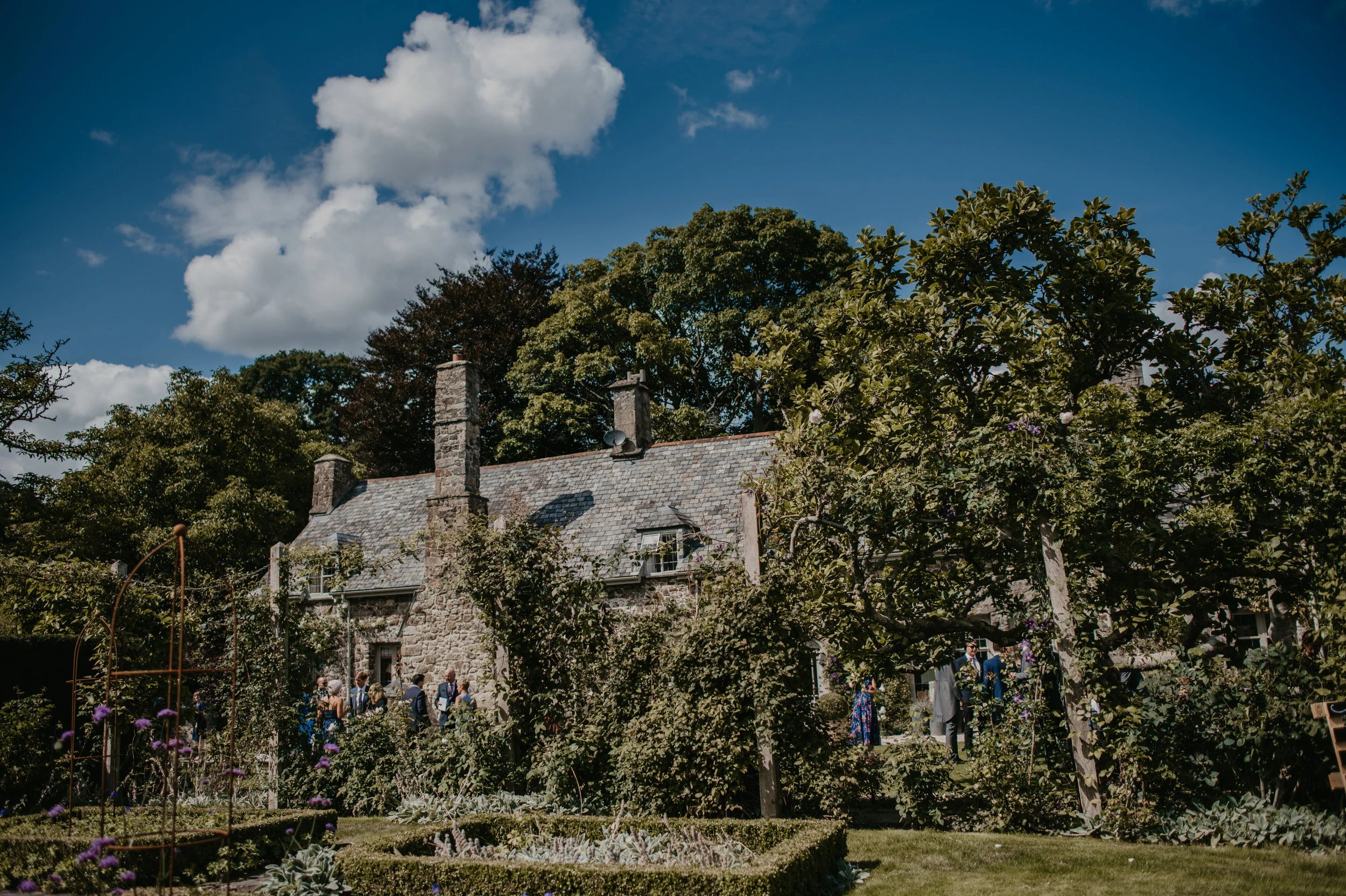 A stone house surrounded by lush greenery and trees, with people gathered outside, possibly for an event, on a bright sunny day with a partly cloudy sky.