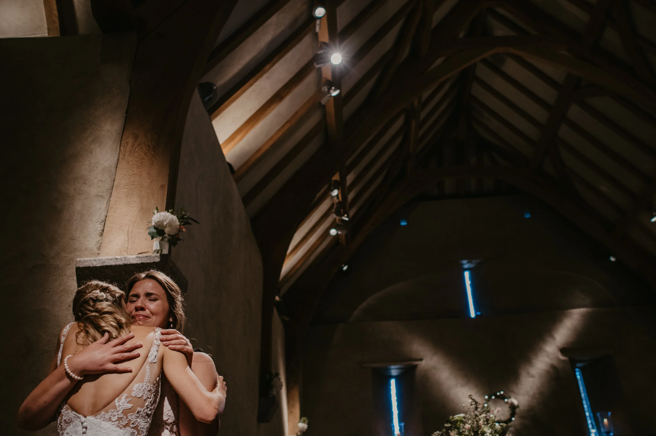 Two women hugging, one in a wedding dress with lace details and the other in a sleeveless dress, in a dimly lit venue with a wooden arched ceiling and floral decorations.
