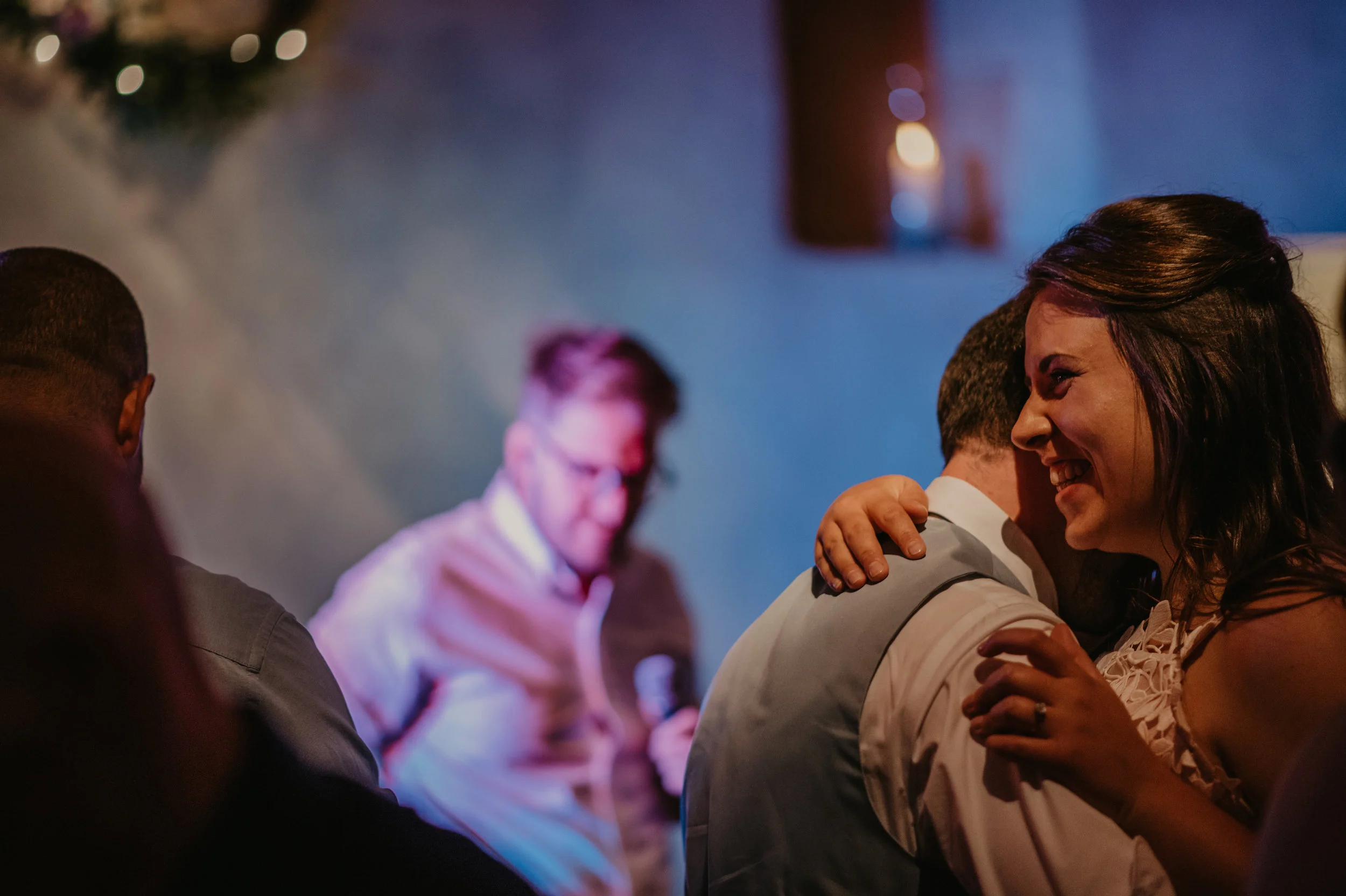 People at a social event, with one woman smiling and hugging a man while others are in the background, in a dimly lit room with blue and warm lighting.