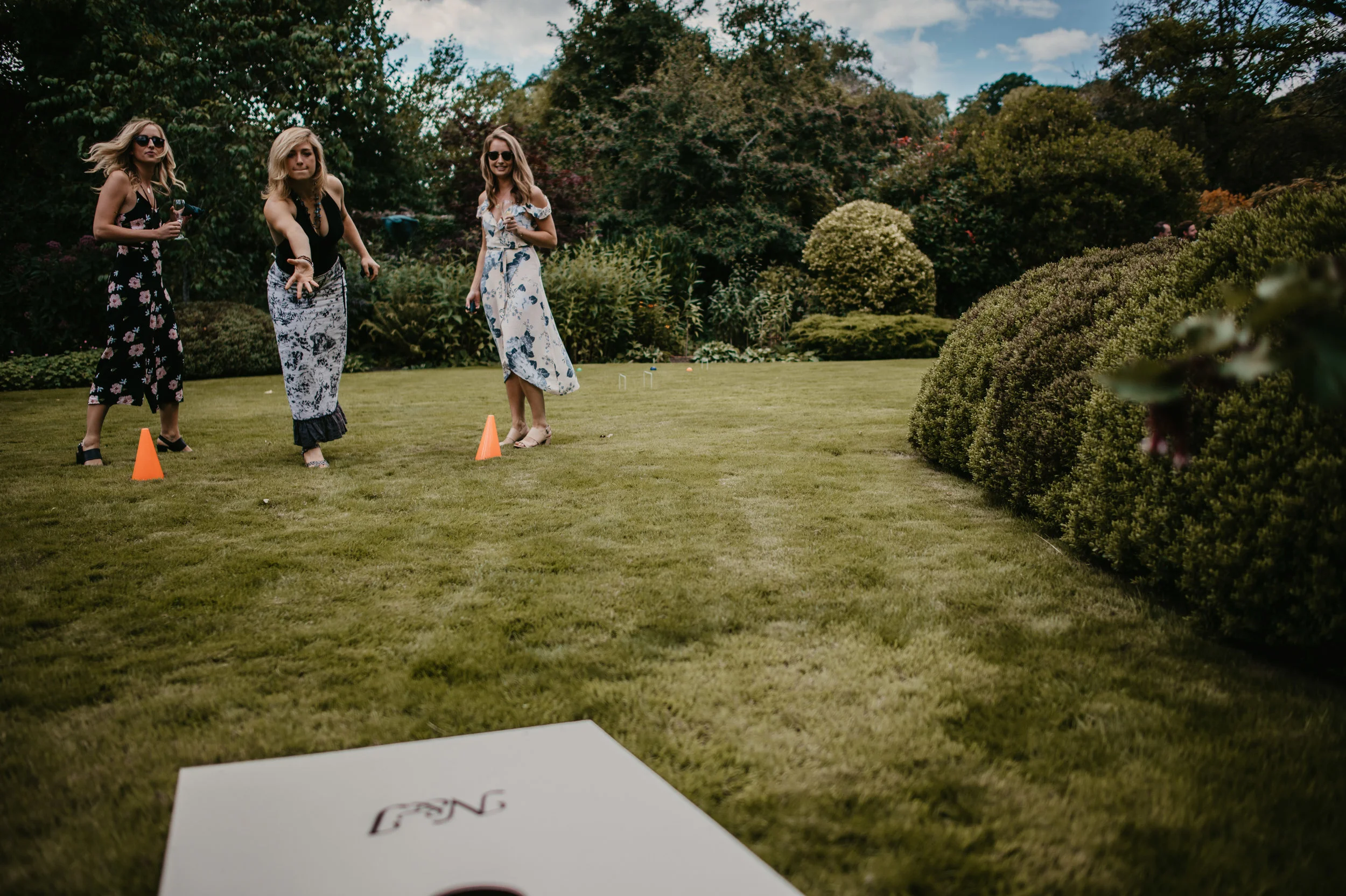 Three women outdoors in a garden, playing cornhole. They are standing near orange cones and dressed in summer clothes, with trees and bushes around them.