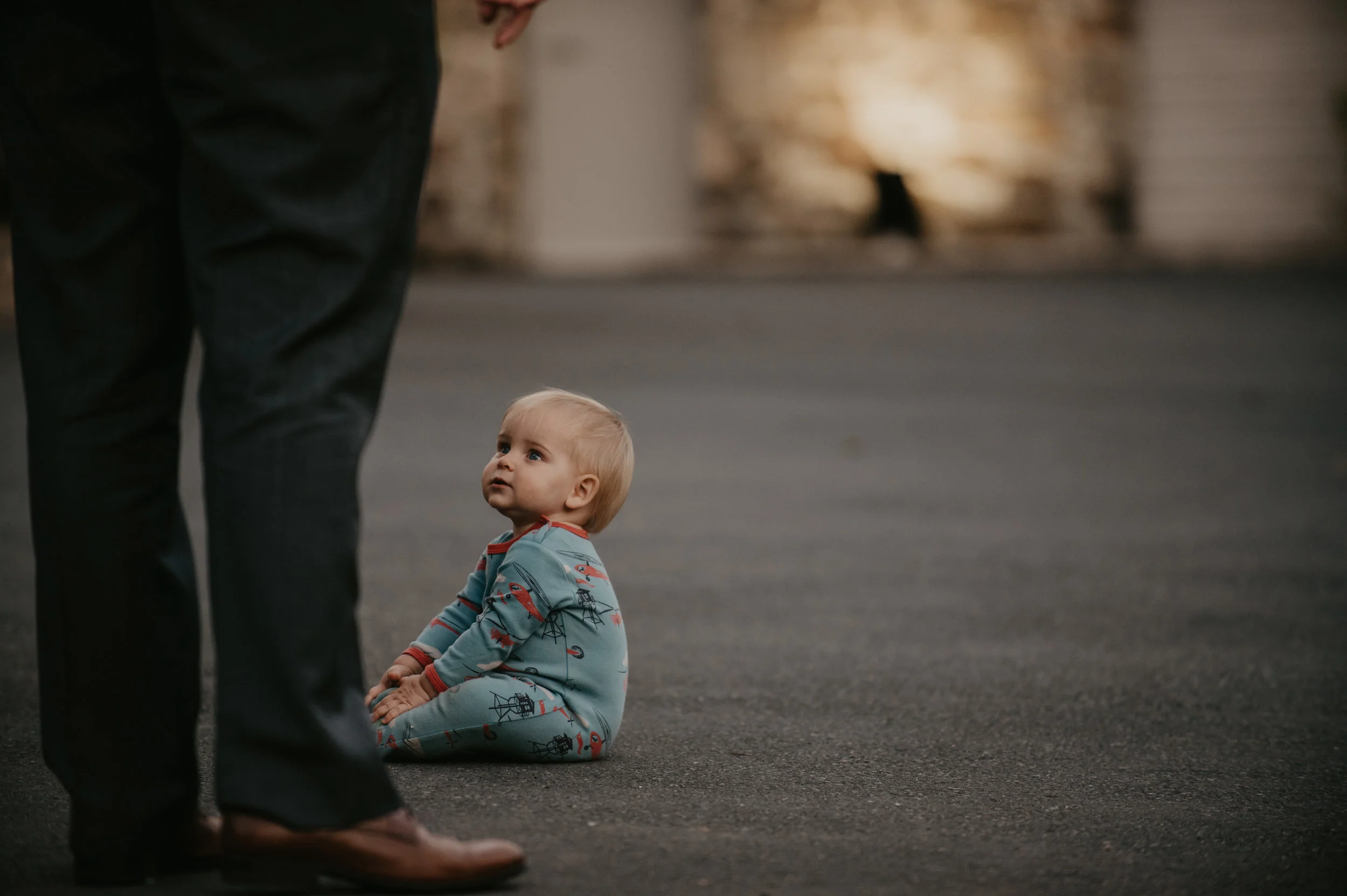 A young child sitting on the ground, looking up with a curious expression, while an adult stands nearby in an urban setting during evening or nighttime.