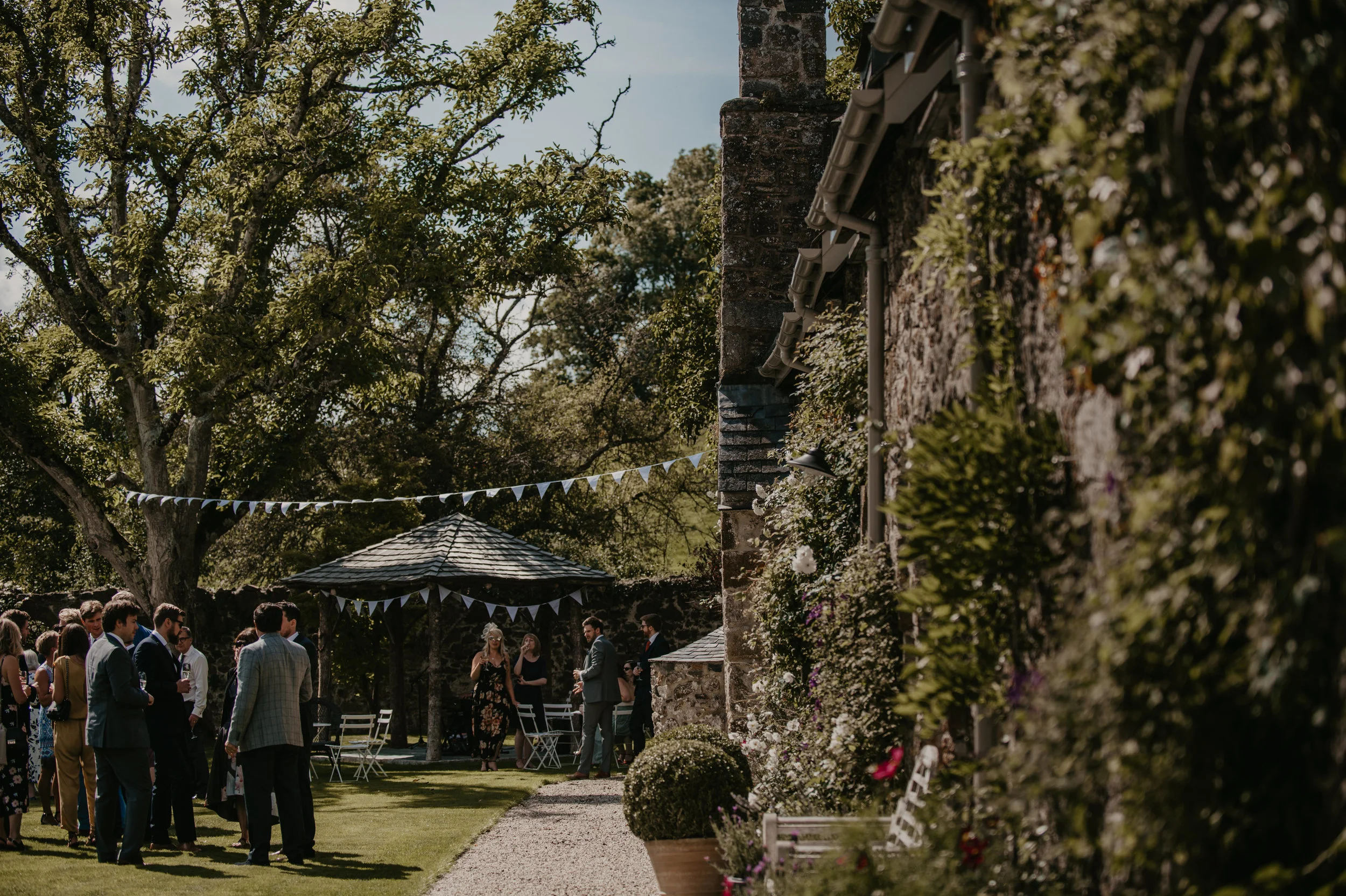 Group of people gathering outdoors during daytime wedding or event with trees, a gazebo, and decorative bunting.