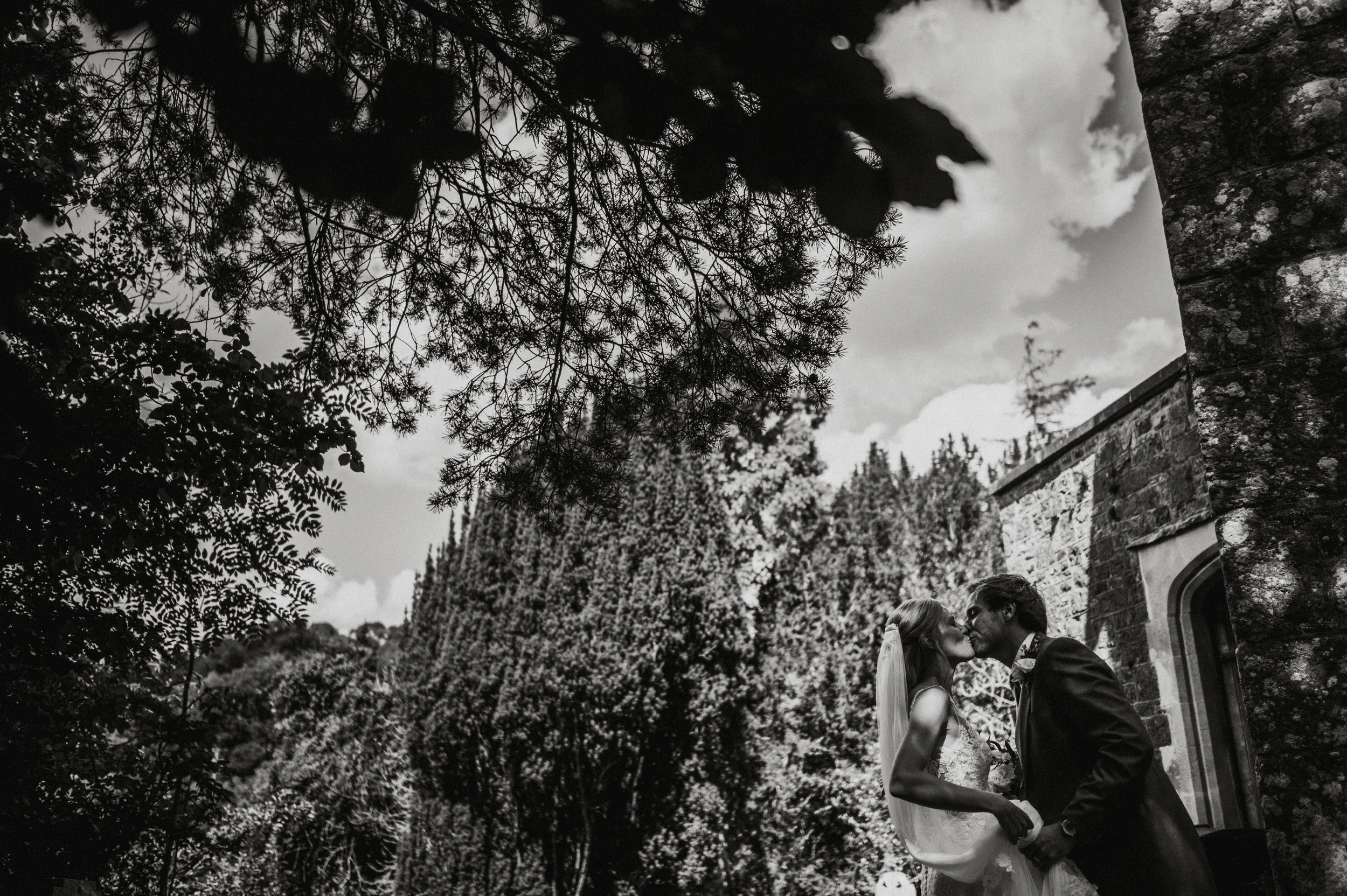 A black and white photograph of a bride and groom sharing a kiss outdoors, with trees and an old stone building in the background.