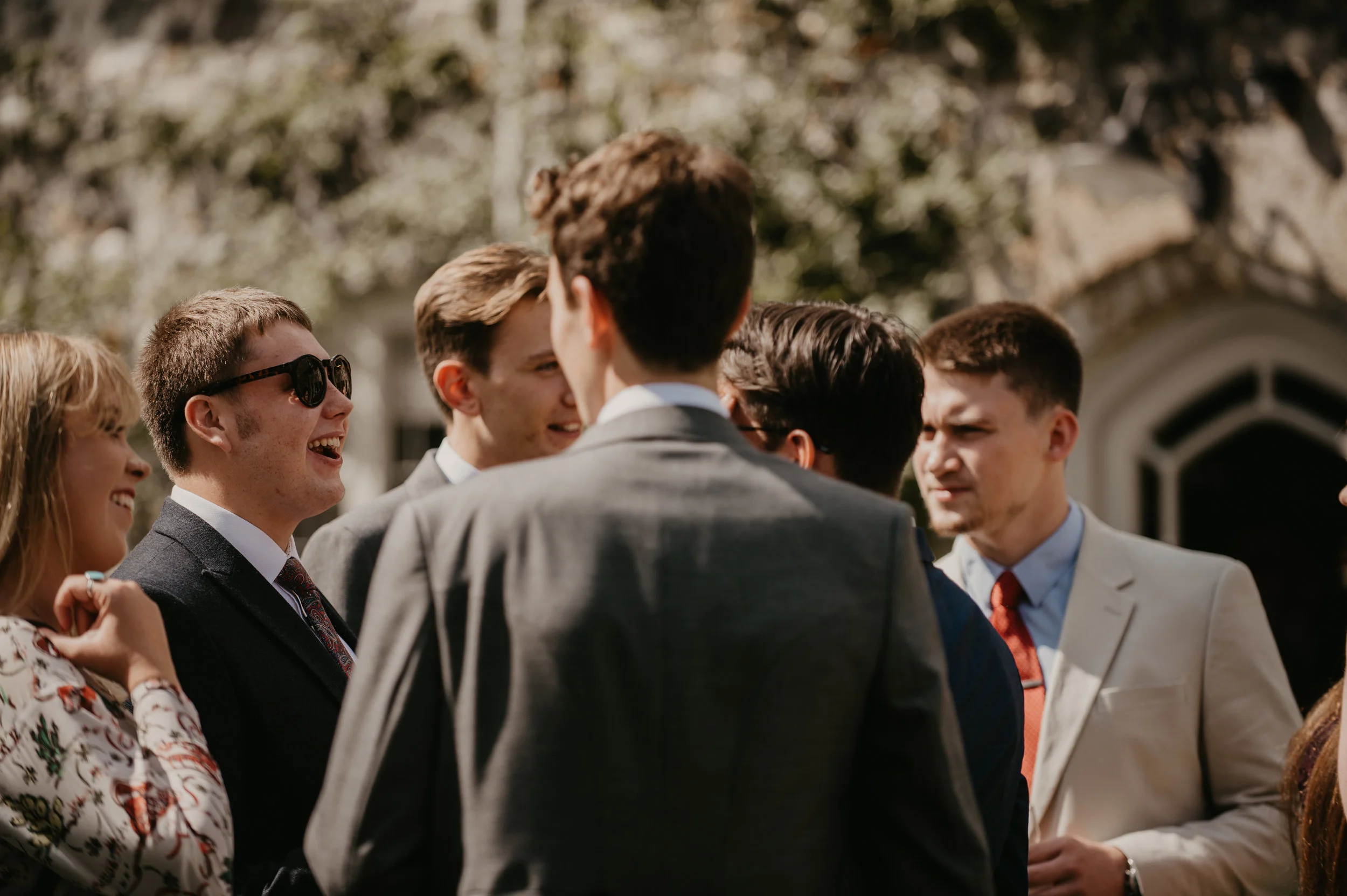 Group of young adults in formal attire smiling and talking outdoors, with a building and trees in the background.