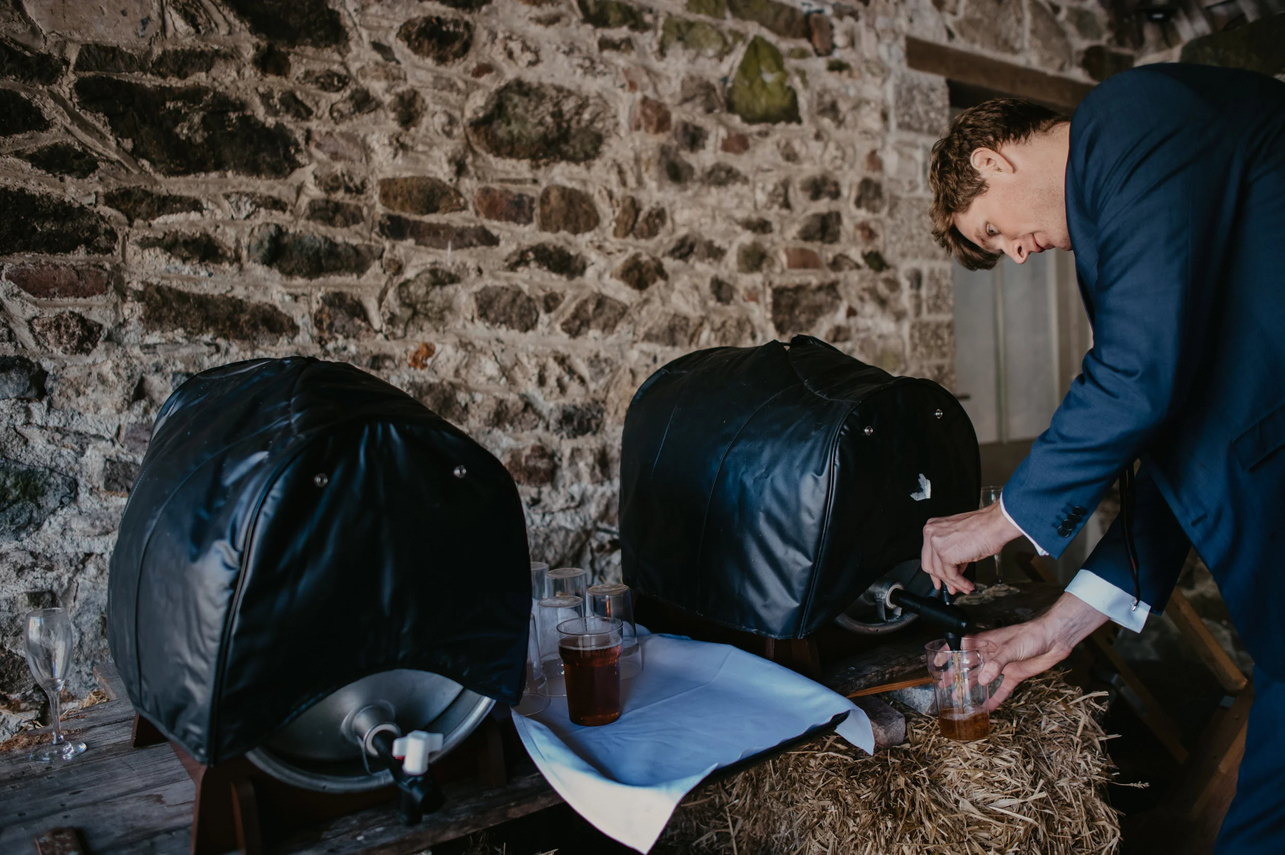 Man in a blue suit pours a drink into a glass from a beer dispenser at a rustic event setup with two black covered beer tanks and glasses on a wooden table.