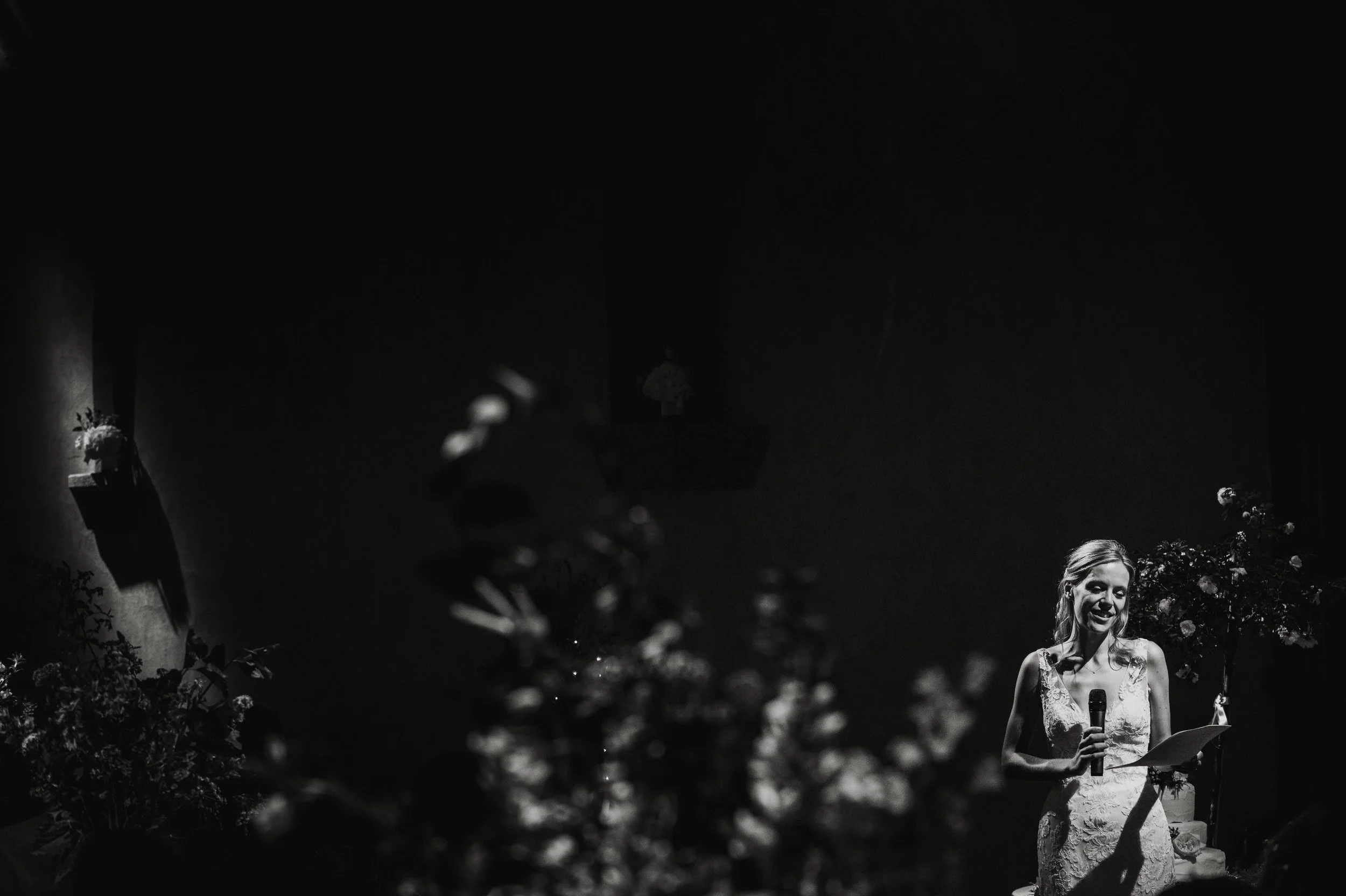 A woman in a wedding dress is giving a speech or reading from notes while holding a microphone, standing in a decorated space with plants and flowers.