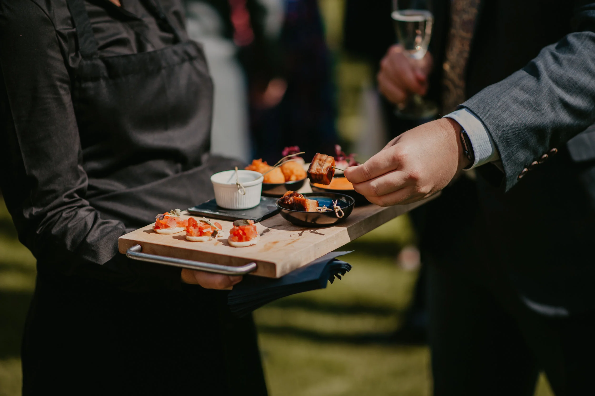 A server in a black apron holds a wooden serving board with small appetizers, including crostini topped with tomatoes and other toppings, and small bowls of sauces or dips. A guest in a suit is using a fork to take a piece of food.