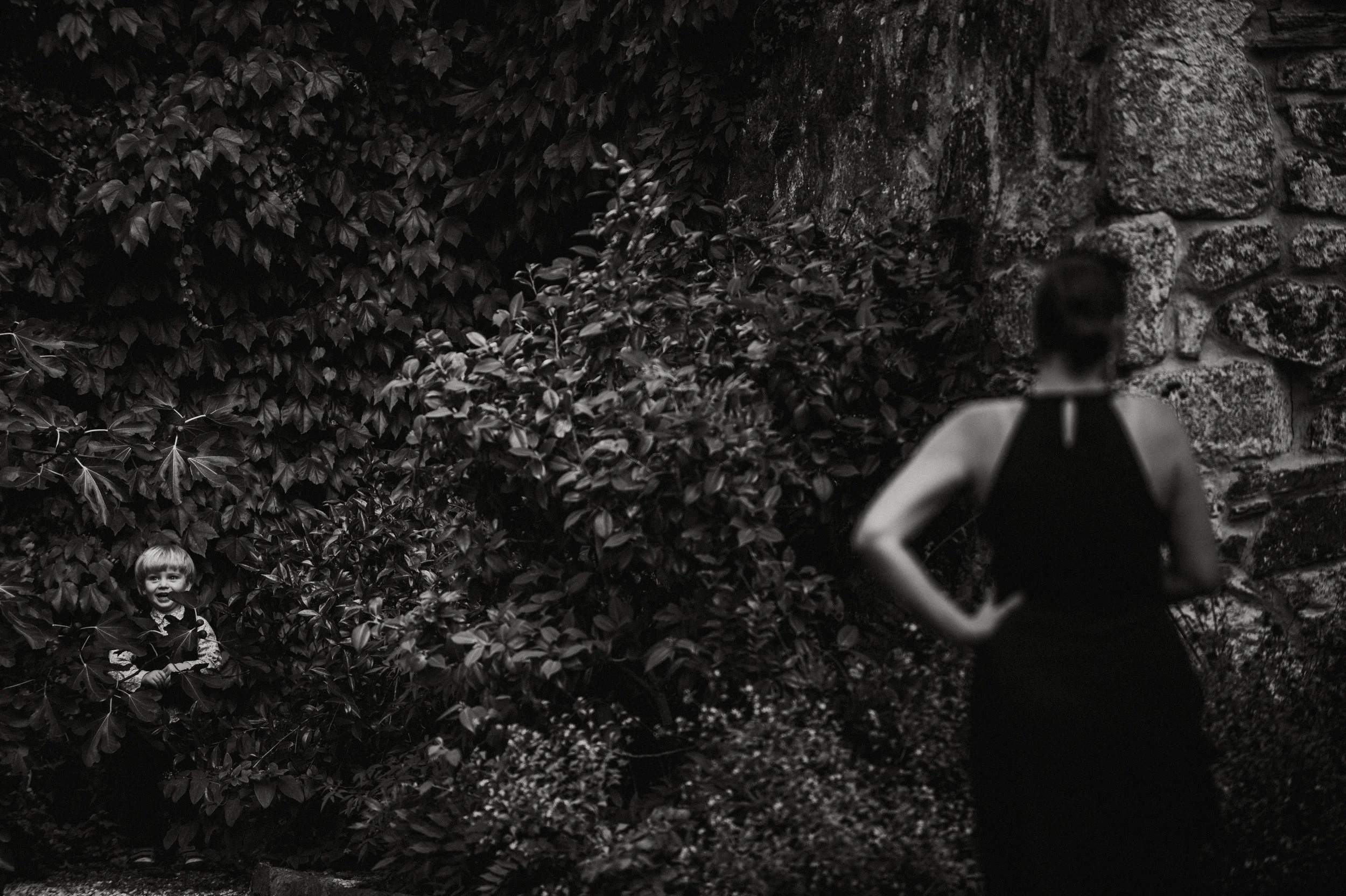 A woman in a black dress with a hand on her hip, standing near a stone wall, looking away. A young boy outdoors among bushes, smiling and holding a stick.