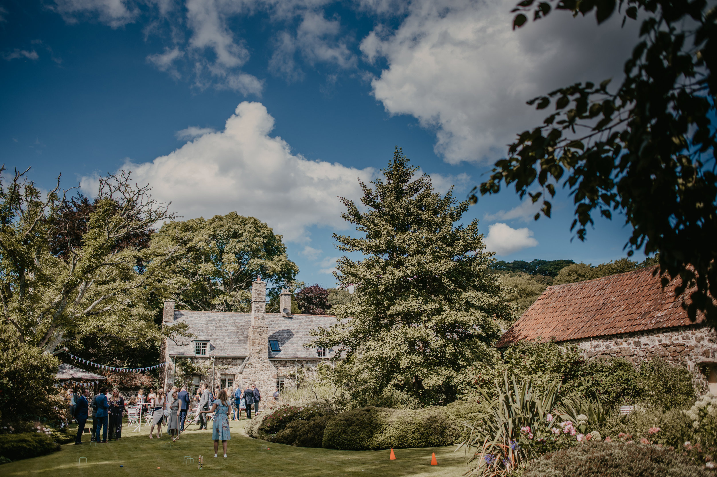 People gathered outdoors on a grassy lawn for a garden party or wedding in a scenic countryside setting with stone cottages, trees, and a blue sky with clouds.