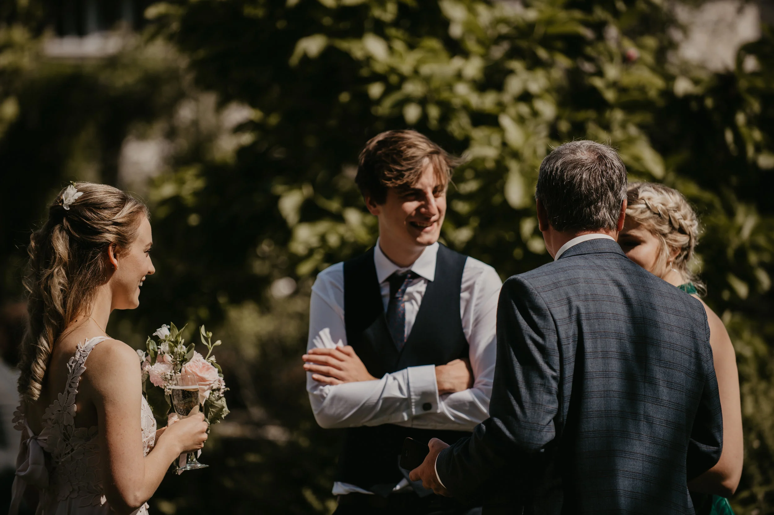 A group of four people, two women and two men, are outdoors engaging in conversation during what appears to be a wedding or formal event, with trees in the background.
