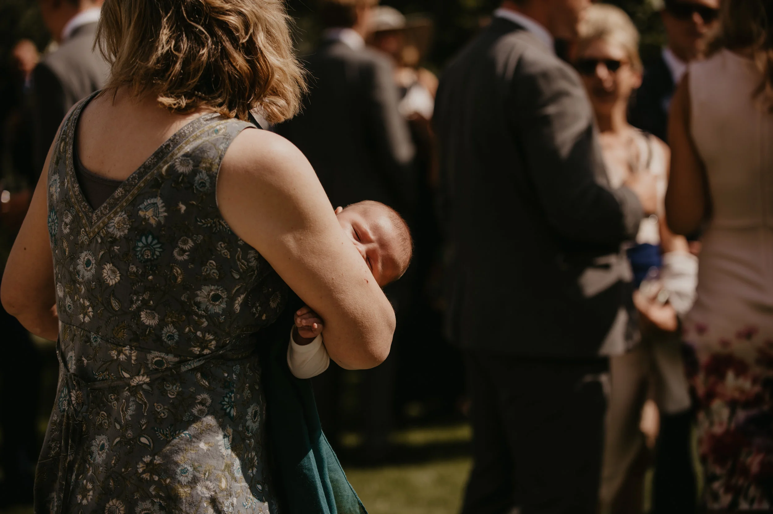A woman holding a sleeping baby during an outdoor gathering with people in formal attire.