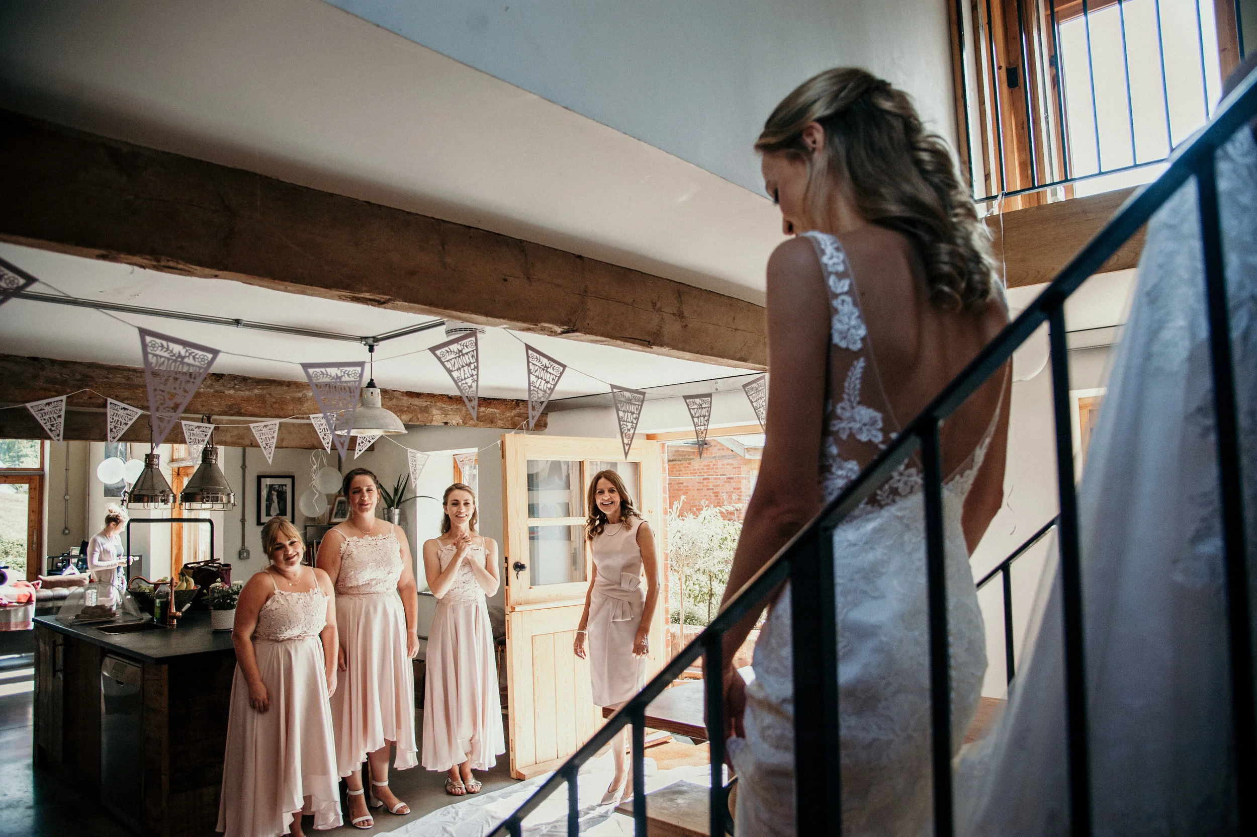 Bridesmaids in matching dresses stand in a room decorated with banners, watching the bride in a lace wedding gown, as she prepares to walk down stairs.