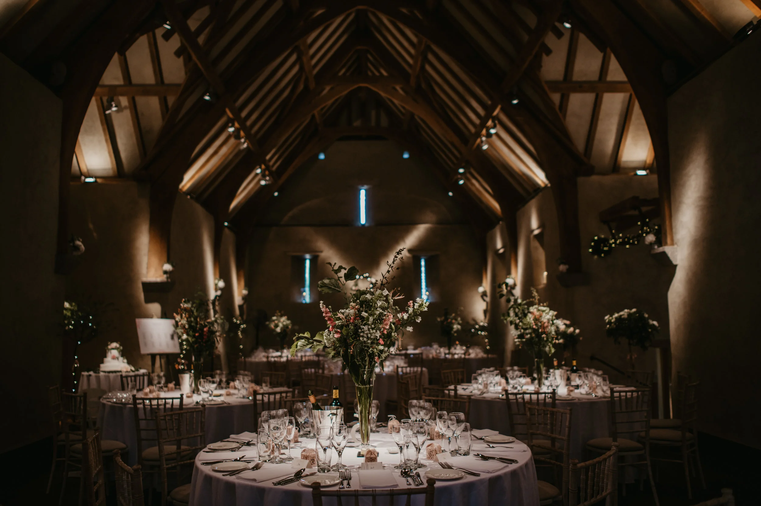 Elegant indoor banquet hall with wooden vaulted ceiling, decorated tables with floral centerpieces, and table settings for a formal event.