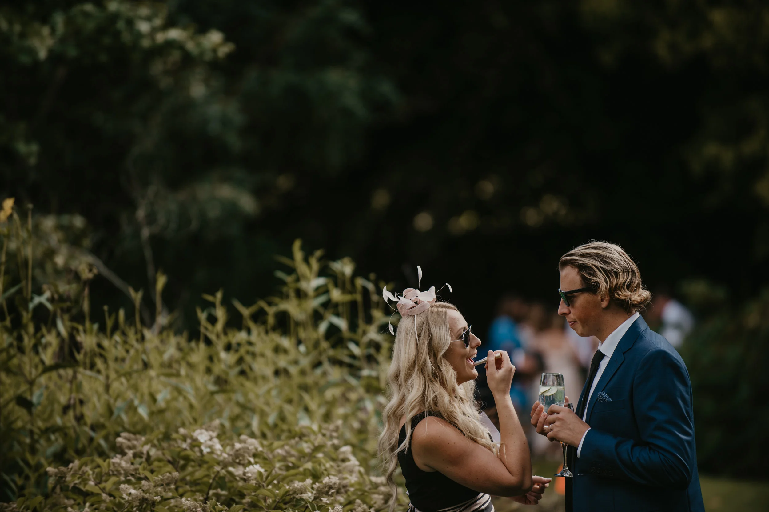 A woman in a fascinator and sunglasses being applied lipstick by a man in a blue suit and sunglasses at an outdoor event, with a background of greenery and other blurred guests.