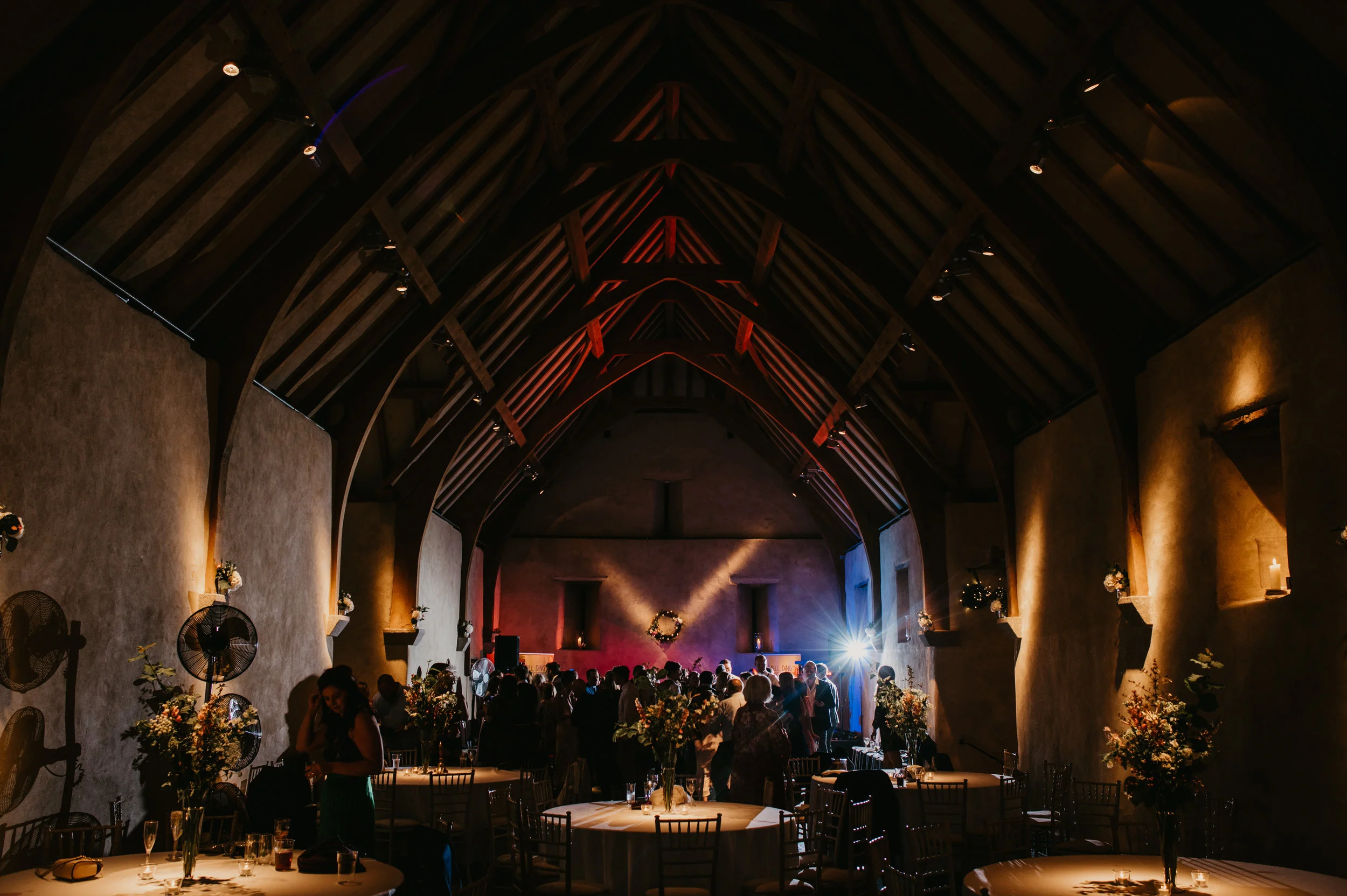 A wedding reception inside a rustic barn with high vaulted wooden ceilings, decorated with flowers on tables, and a dance floor illuminated by colorful lights, with guests mingling and enjoying the event.