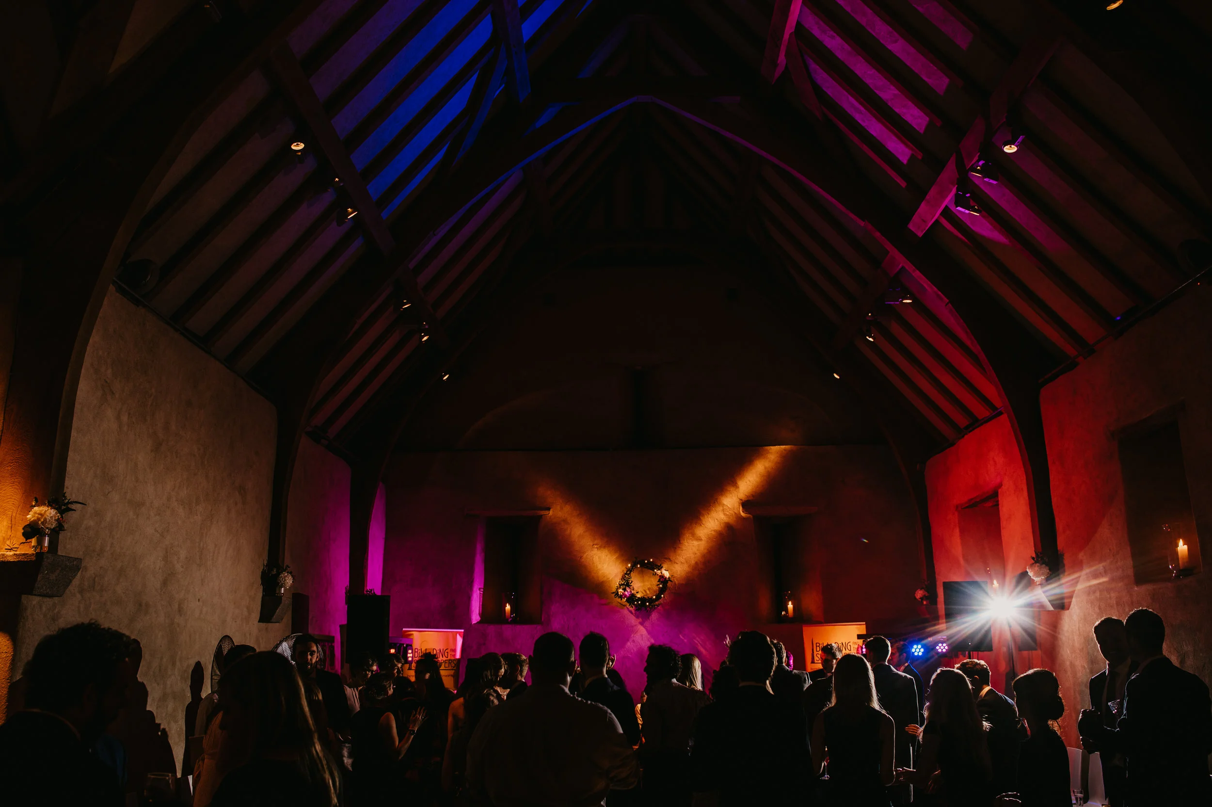 People gathered in a dimly lit, decorated hall with high, wooden ceiling, colorful lighting, and candles for a celebration or event.