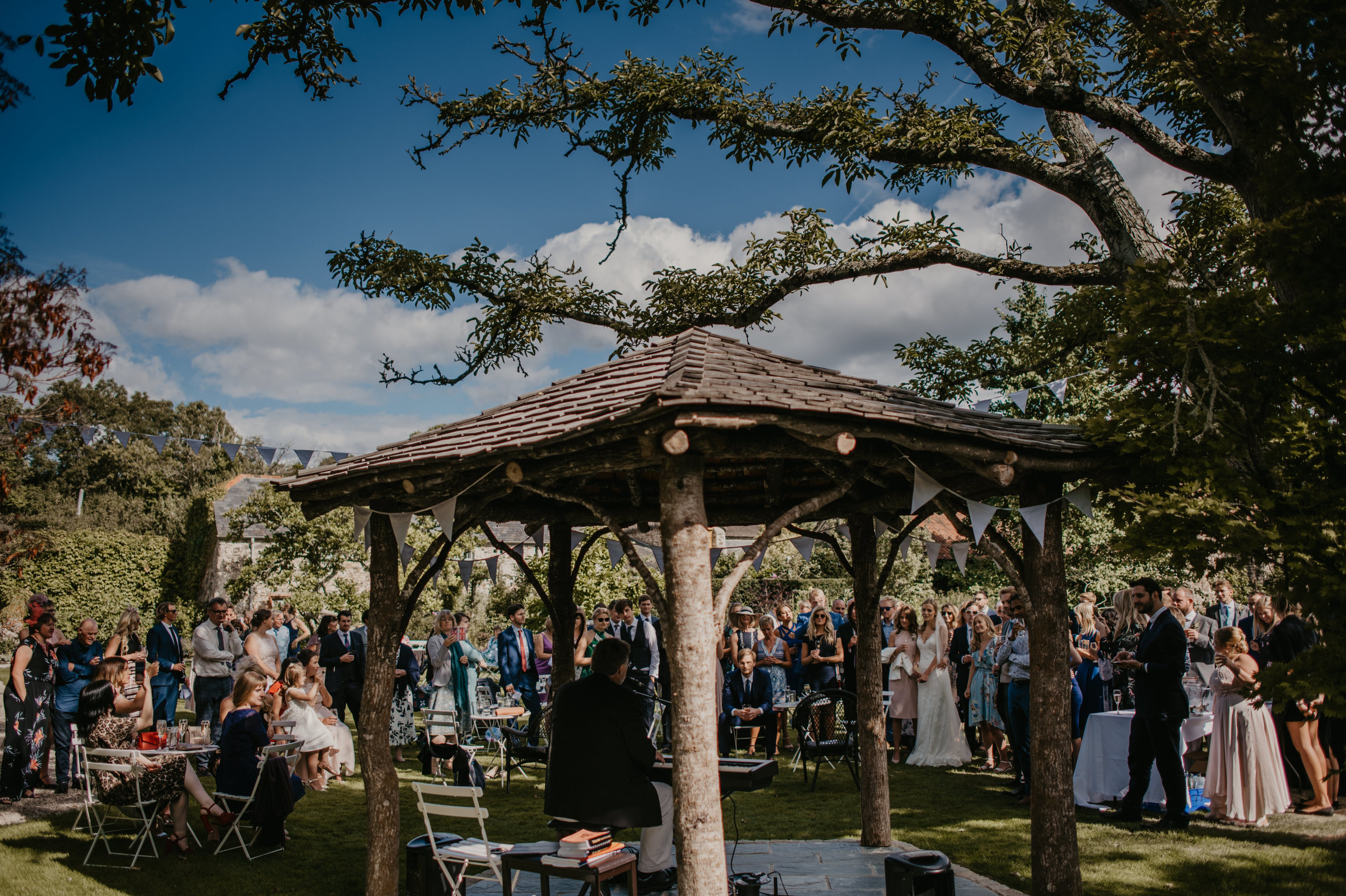 Outdoor wedding reception in a garden with many guests, a singer, and a pianist performing under a shelter, surrounded by trees and party decorations.
