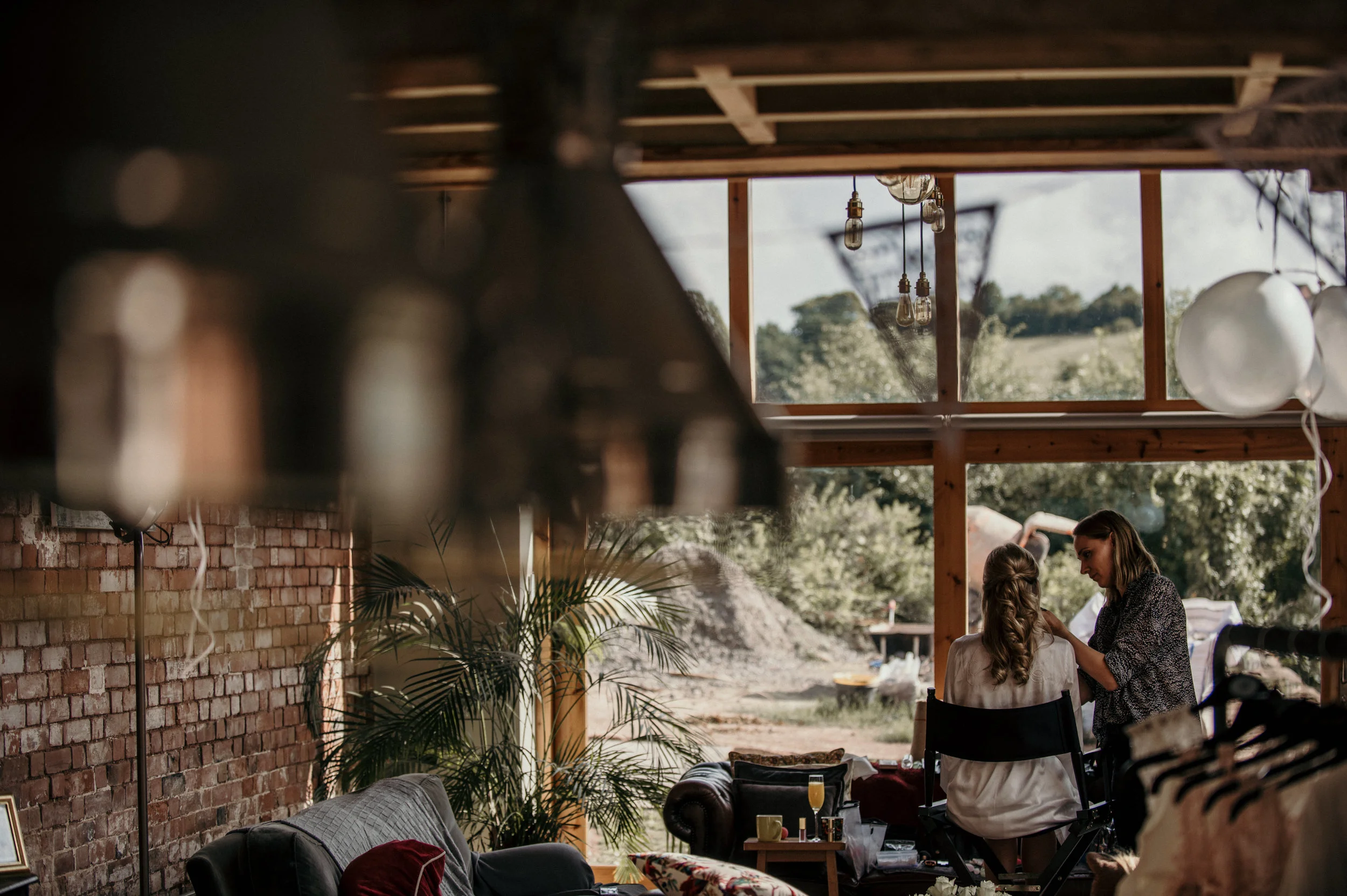 A woman getting her makeup done by another woman inside a rustic, natural-light studio with exposed brick wall, a large window overlooking a garden, and various decorative elements like hanging bulbs, white balloons, and indoor plants.