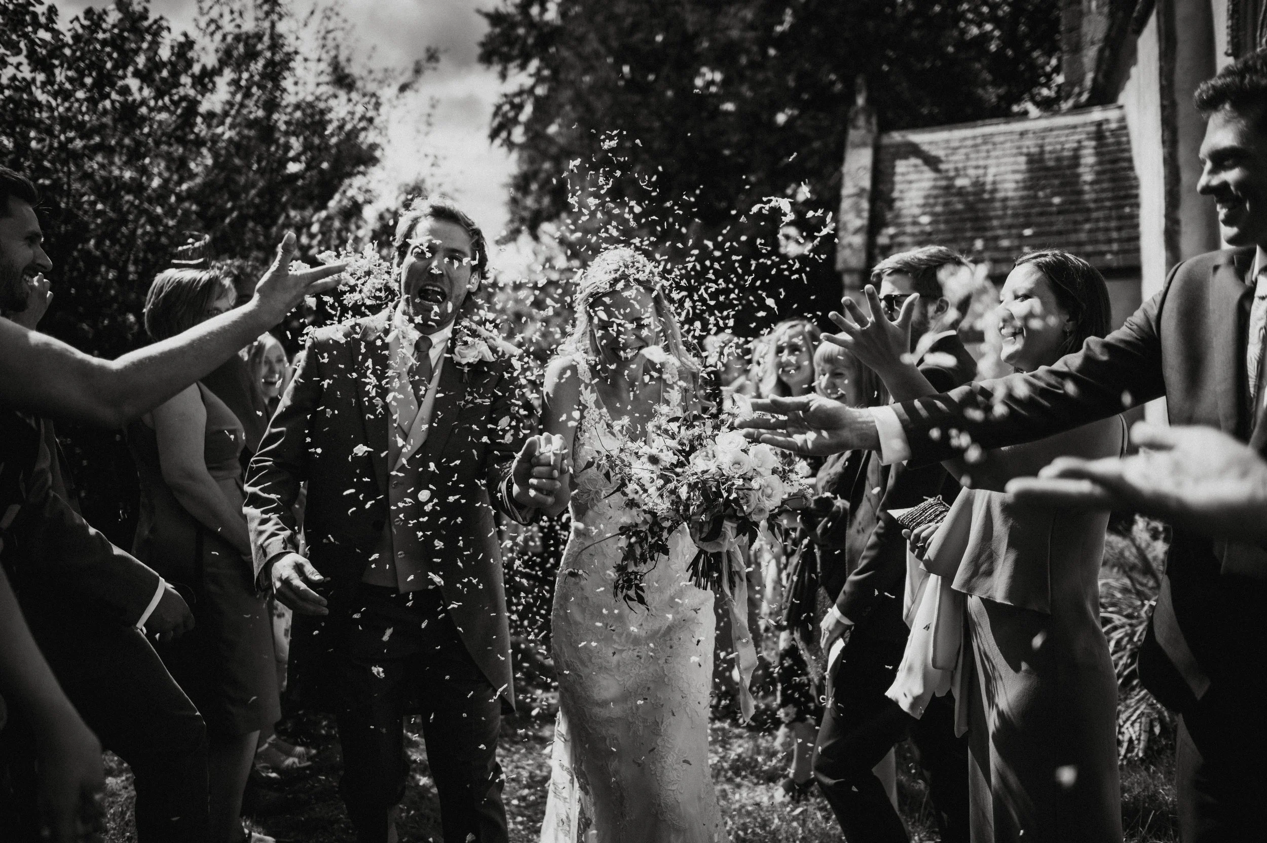 Black and white photo of a wedding celebration with the bride and groom walking through a crowd of guests throwing confetti at them outdoors.